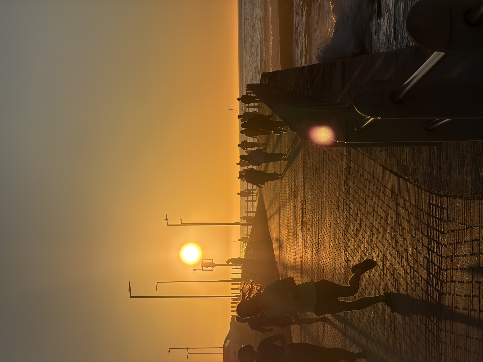 The image depicts a sunrise over the ocean in Cape Town, South Africa, likely along the Sea Point promenade. Silhouettes of people are visible, engaged in various activities such as running and walking along the paved walkway. The sun is low on the horizon, casting a warm, golden light and long shadows across the path. Streetlights are spaced along the promenade. The ocean is to the right of the walkway, with gentle waves breaking on the shore. In the distance, a sailboat is visible on the water. The overall atmosphere is serene and active, capturing a typical morning scene in this coastal city.