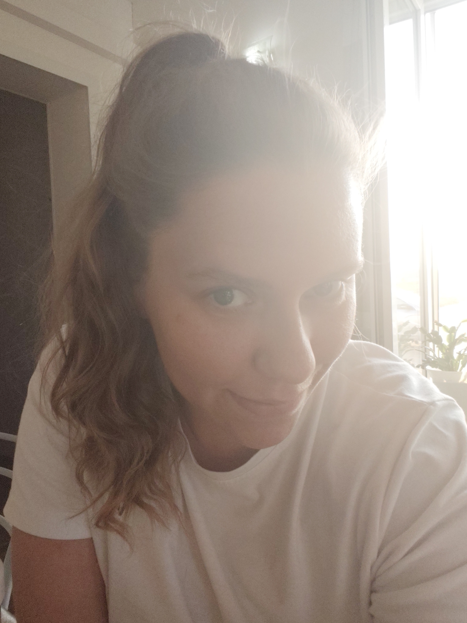 A woman is facing the camera. She has her blonde hair partially up in a bun and a white t-shirt. The background includes a door, window, and a plant. The image appears to be taken indoors in the city of Нижний Пальник, Russia.