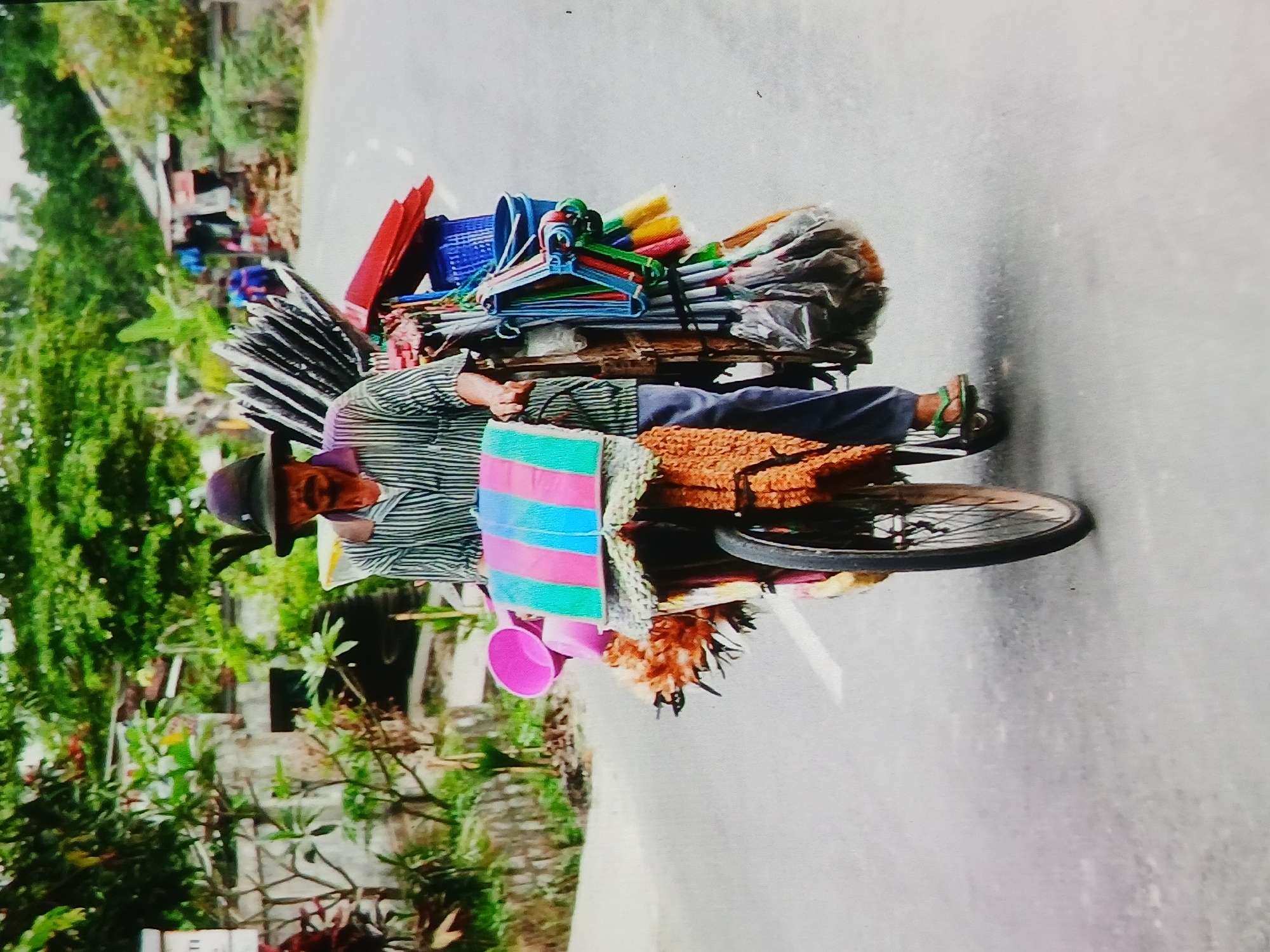 A man with a dark, wide-brimmed hat rides a bicycle laden with various items. He wears a striped shirt and blue pants. The bicycle is heavily loaded with brooms, brushes, baskets, and other items, some wrapped in plastic. A pink bucket is attached to the front of the bicycle, along with a colorful striped cloth cover. The man is on a paved road, with greenery and buildings visible in the background. The scene appears to be in a village or rural setting within Setia Bakti, Indonesia.