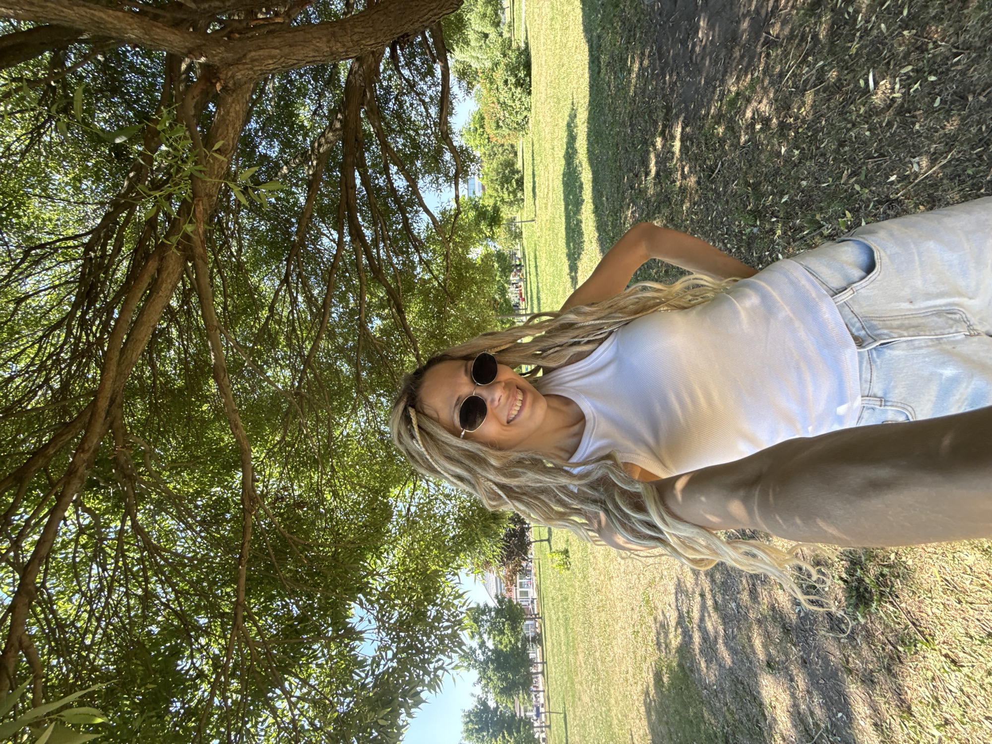 A young woman with blonde dreadlocks, wearing a white tank top, sunglasses, and light-wash denim shorts, is taking a selfie in a park in the city of Zavolzhye, Russia. She is smiling and standing under a large tree with green leaves, with dappled sunlight filtering through the branches. The background shows a grassy field with other trees and bushes, and some buildings in the distance. The overall scene suggests a pleasant day outdoors in a park setting.