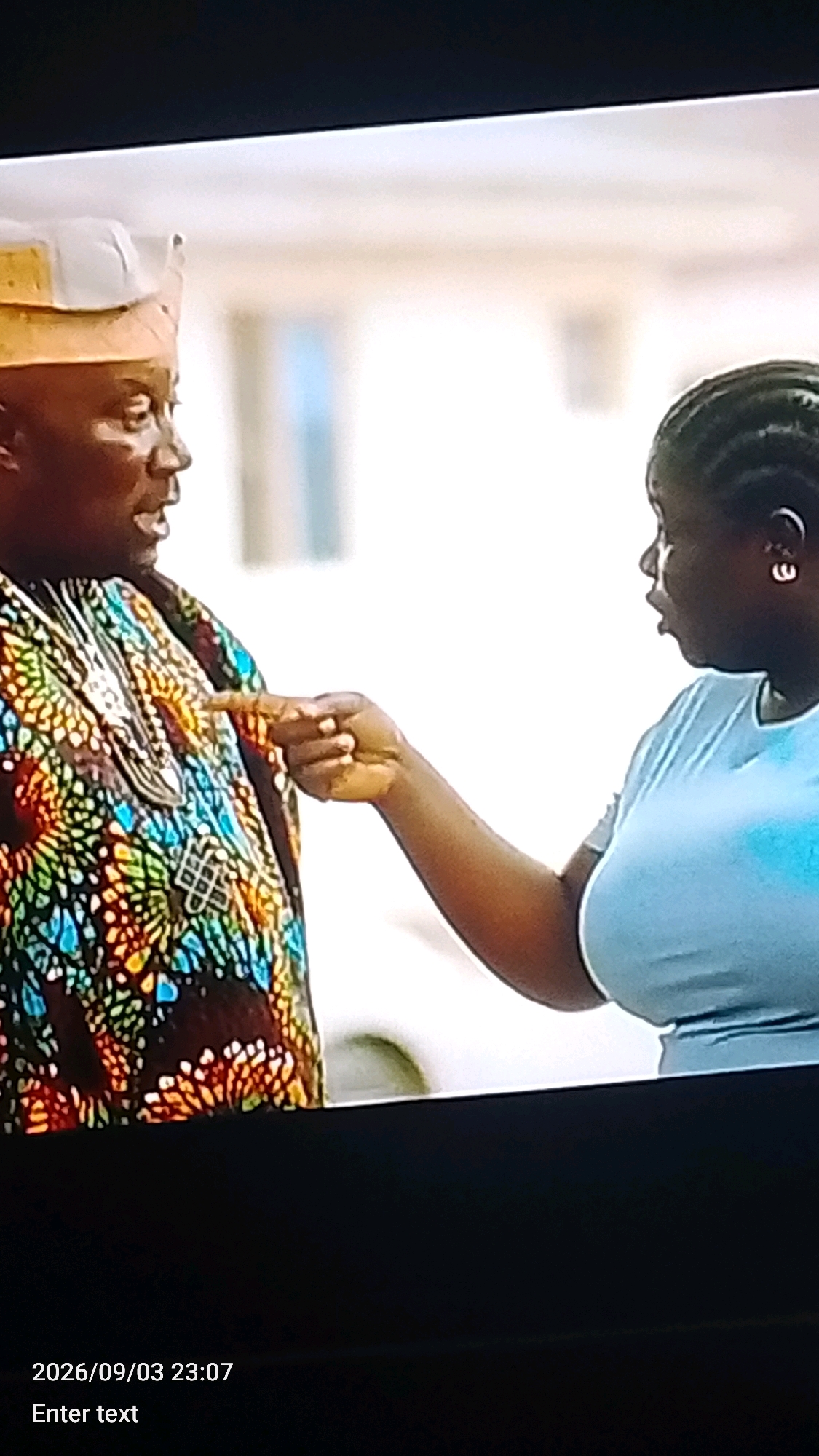 A man wearing a traditional yellow and white cap and a colorful patterned garment with ornate silver jewelry is speaking with a woman wearing a light blue t-shirt. The woman is gesturing with her right hand towards the man, and they appear to be having an intense conversation. The background is blurred, suggesting an outdoor urban setting, possibly in Lokoja, Nigeria, with hints of light-colored buildings. The image is a screenshot from a video, as indicated by the date and time stamp "2026/09/03 23:07" and the text "Enter text" at the bottom.