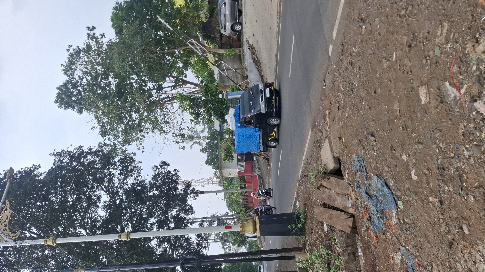 The image captures a street scene in Cilaku, Indonesia, characterized by a road, roadside debris, and vegetation. A black Suzuki carry truck, covered with a blue tarp and carrying cargo, is the primary vehicle on the paved road, driving towards the right. Two motorcyclists, one with a passenger, are also on the road, traveling in the same direction as the truck. To the left of the road, a white lamppost with ornate gold accents stands prominently. The roadside on the left is bordered by a ditch with wooden planks and scattered debris, including plastic sheeting and general litter. The right side of the road features a slight incline with a retaining wall, more vegetation, and a grey SUV parked further down. Buildings, including what appears to be a house with a red and white facade and a separate structure with a pitched roof, are visible in the background, surrounded by trees and foliage. A telecommunications tower is also visible in the distance. The sky is overcast.