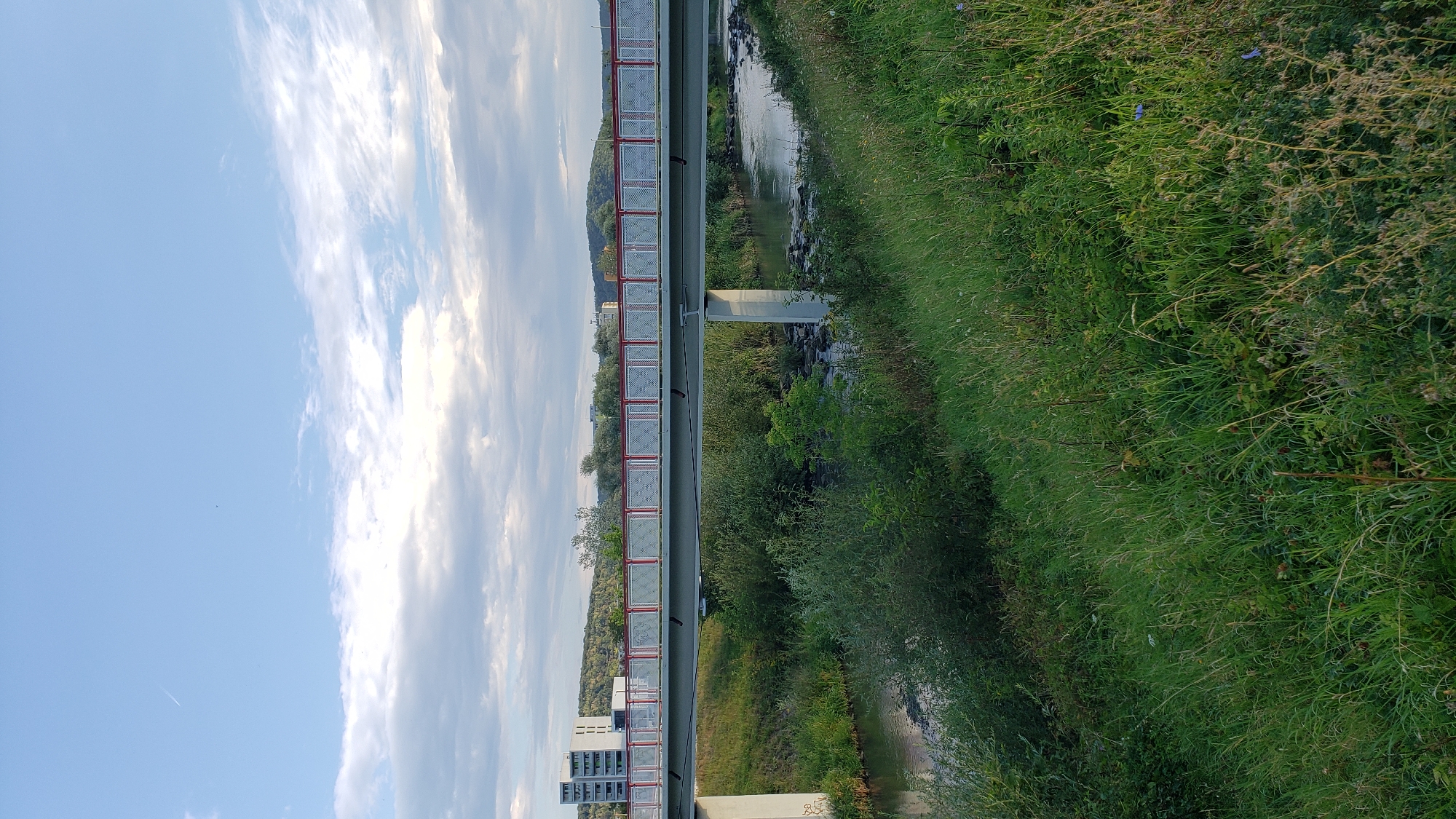 The image depicts a pedestrian bridge spanning a small river in Prešov, Slovakia. The bridge has a gray deck with red railings and a white mesh-like railing infill. The river flows through a green, overgrown bank on the right side of the image, with rocks visible in the water. On the left bank, there is a steep, grassy embankment leading up to the bridge, with trees and bushes also present. In the background, on the left, a tall, modern building with several floors is visible, along with more trees and hills. The sky is partly cloudy with a light blue hue. The overall scene appears to be a calm, natural setting within or on the outskirts of a city. There are no people or animals visible in the image.