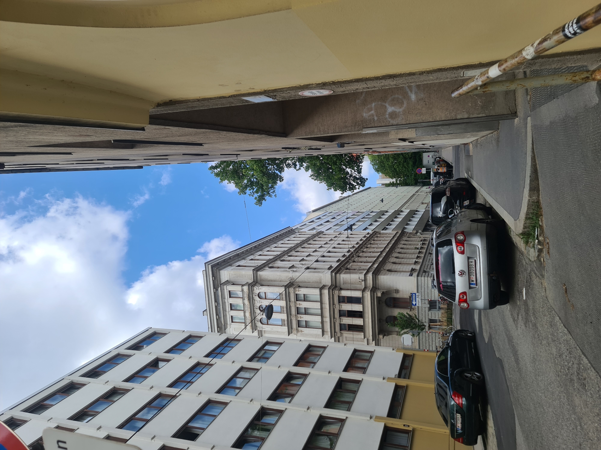 The image depicts a street scene in Vienna, Austria, characterized by a row of buildings and parked cars. On the left side of the street, a modern, white apartment building with numerous windows stretches upwards. Adjacent to it, a more ornate, older building with classical architectural details, including arched windows and decorative elements, stands tall. The street itself is paved, with a sidewalk running alongside the buildings. Several cars are parked along the street, including a dark green sedan and a silver station wagon. The sky above is blue with scattered white clouds, and trees are visible in the background, suggesting a leafy urban environment. The perspective of the photograph is from street level, looking down the road.