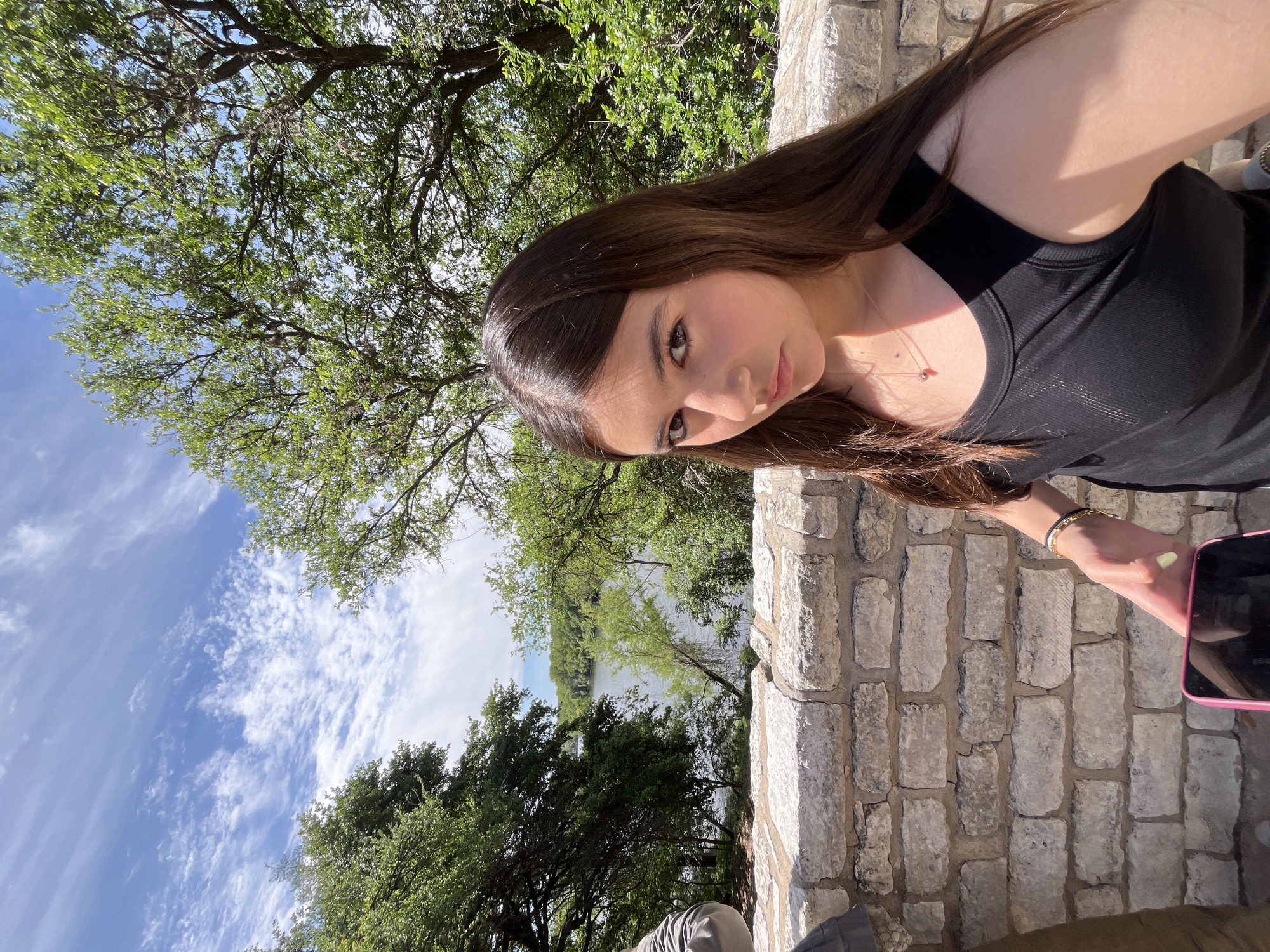 A young woman with long brown hair and wearing a black tank top stands next to a stone wall. She is looking at the camera. The background includes trees, a body of water, and a sky with clouds. Based on the landscape and stonework, it is likely that the location is along the Ann and Roy Butler Hike-and-Bike Trail at Lady Bird Lake in Austin, Texas.