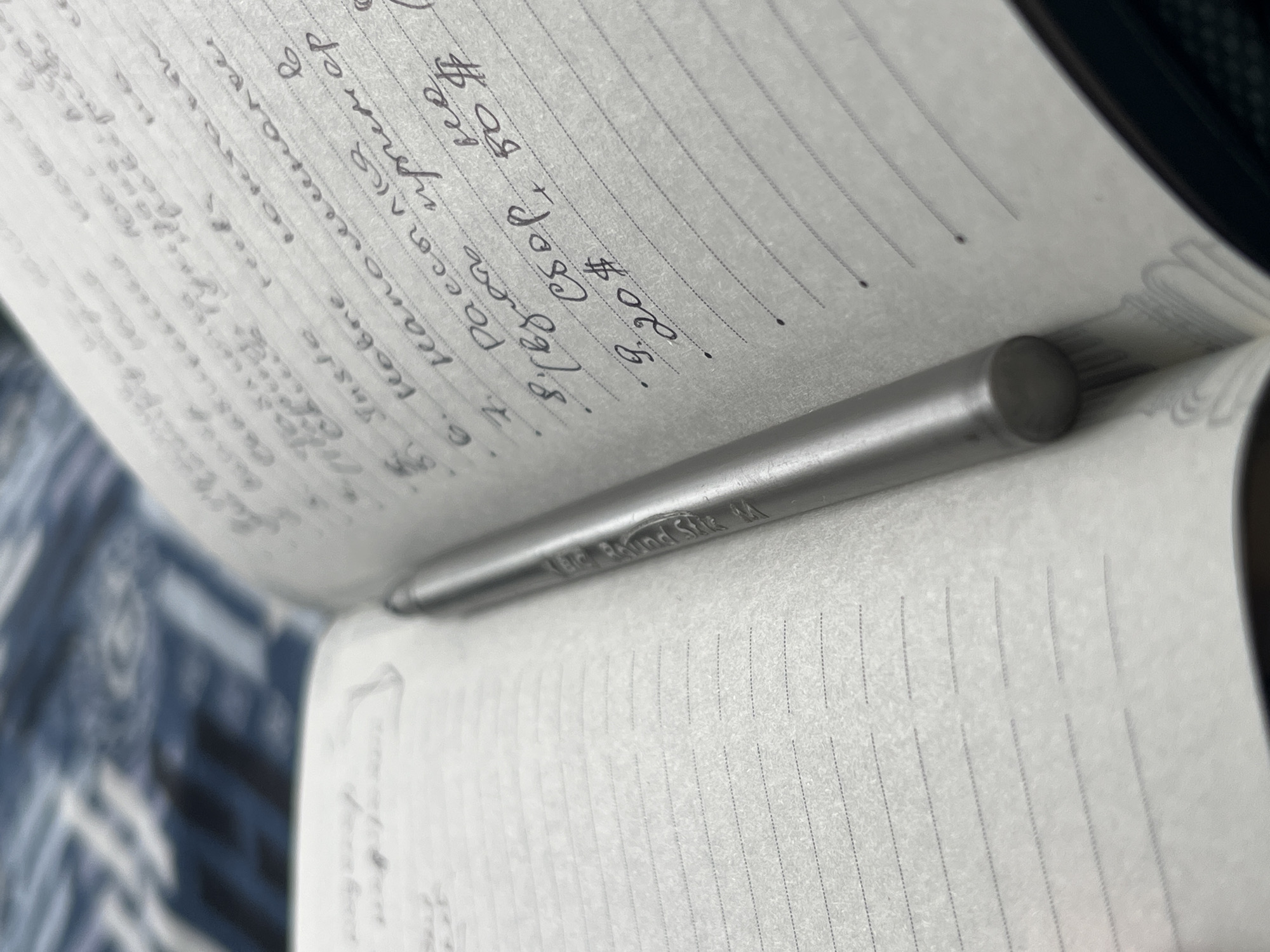 A metallic pen rests on an open notebook with lined pages. The notebook is filled with handwritten text and a numbered list in black ink. The background is a blurry, dark blue and white patterned fabric. The image does not contain any information about the location in the city of ღრმაღელის თემი, Georgia.