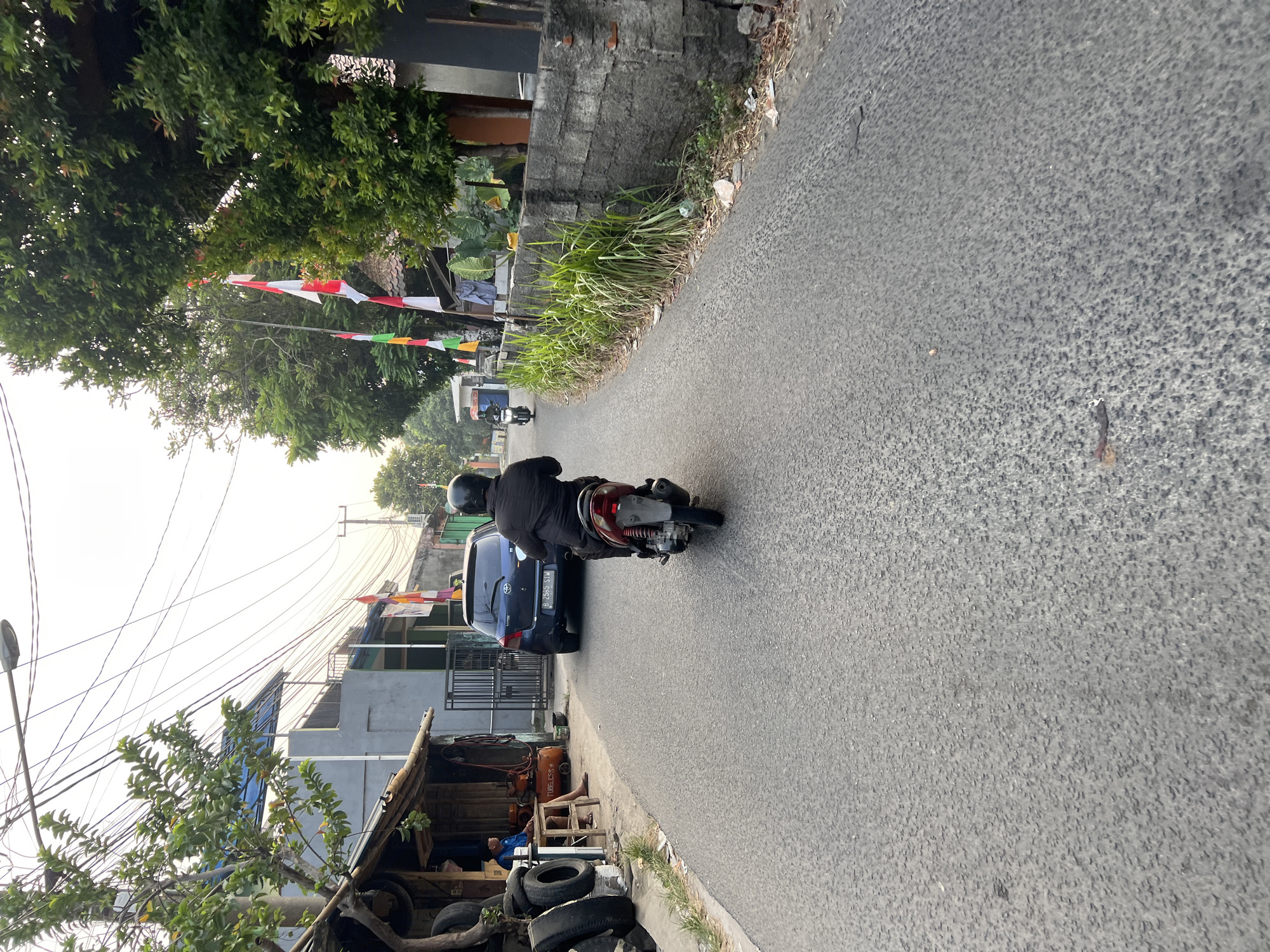 A person wearing a black jacket and black helmet is riding a red and silver scooter on a paved road in Klapanunggal, Indonesia. The scooter is moving away from the viewer. In the background, a dark blue car is parked on the left side of the road. Further down the road, another scooter is visible. Buildings and trees line the right side of the road, with Indonesian flags attached to poles. Electrical wires are strung across the sky. The environment appears to be a residential or commercial area in a town or city.