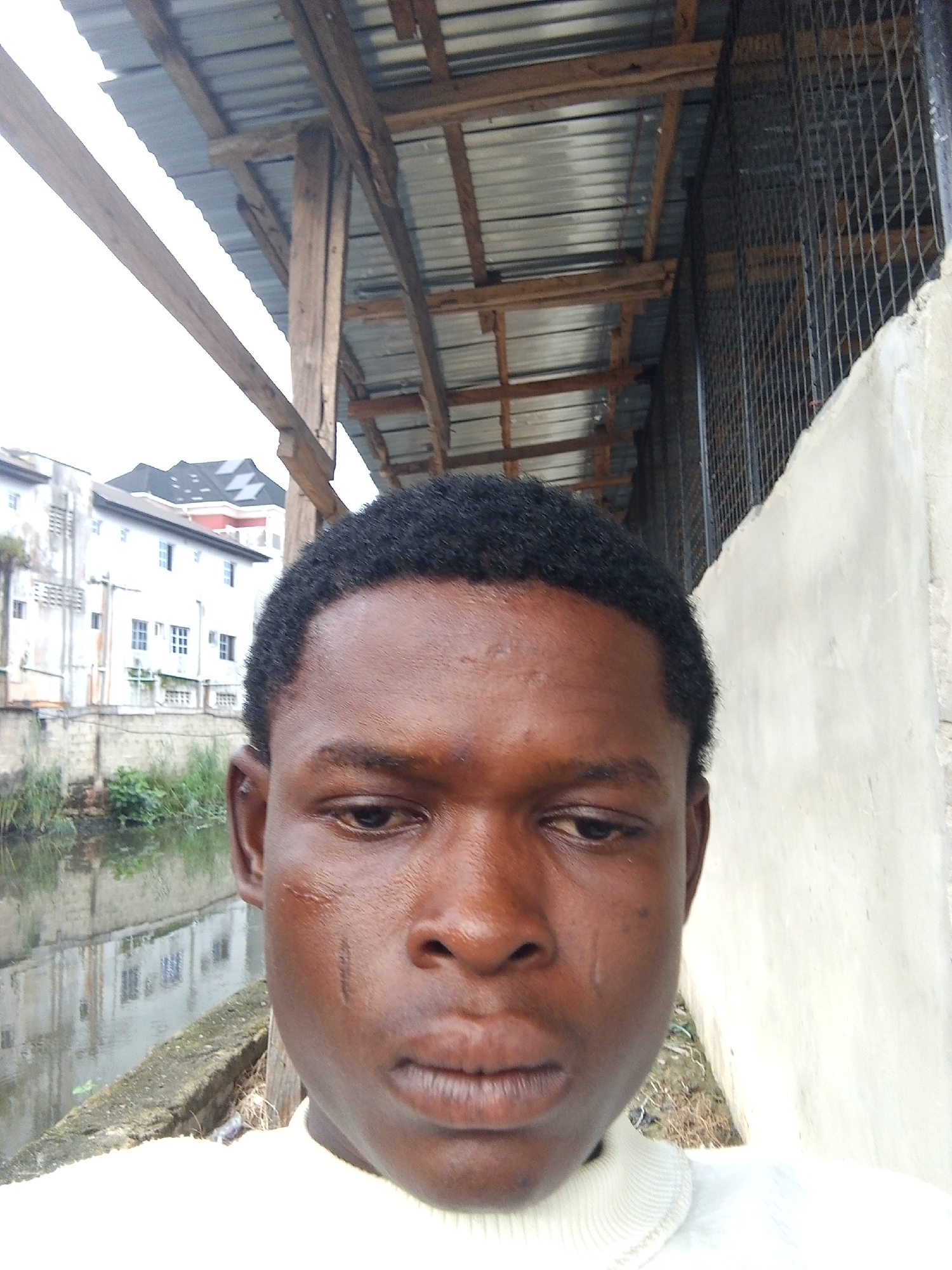 The image captures a close-up selfie of a young Black man, centered prominently in the foreground. He has short, dark, curly hair and is wearing a light-colored, possibly off-white, turtleneck top. Distinctive features on his face include several dark scarification marks on his left cheek and forehead. A faint streaked mark on his right cheek might suggest a tear. His eyes are cast downwards, conveying a somber or contemplative expression.
The background unfolds an urban or semi-urban outdoor scene. To the man's left, a body of water, resembling a canal or river, flows alongside a concrete bank with some scattered green vegetation. Across this waterway, several light-colored, multi-story buildings with various windows are visible, one featuring a dark, intricate roof design.
Above the man and to his right, a makeshift shelter or canopy is constructed from raw wooden beams and posts, supporting a corrugated metal roof. Further to the right, a plain, light-colored concrete wall is visible, with a dark, grid-like metal fence structure behind it.
The lighting is soft and diffused, indicating an overcast or cloudy daytime. The mix of residential buildings, a waterway, and simple infrastructure aligns with typical scenes in Ado, Nigeria, which is provided as the location context. No text is visible in the media.
