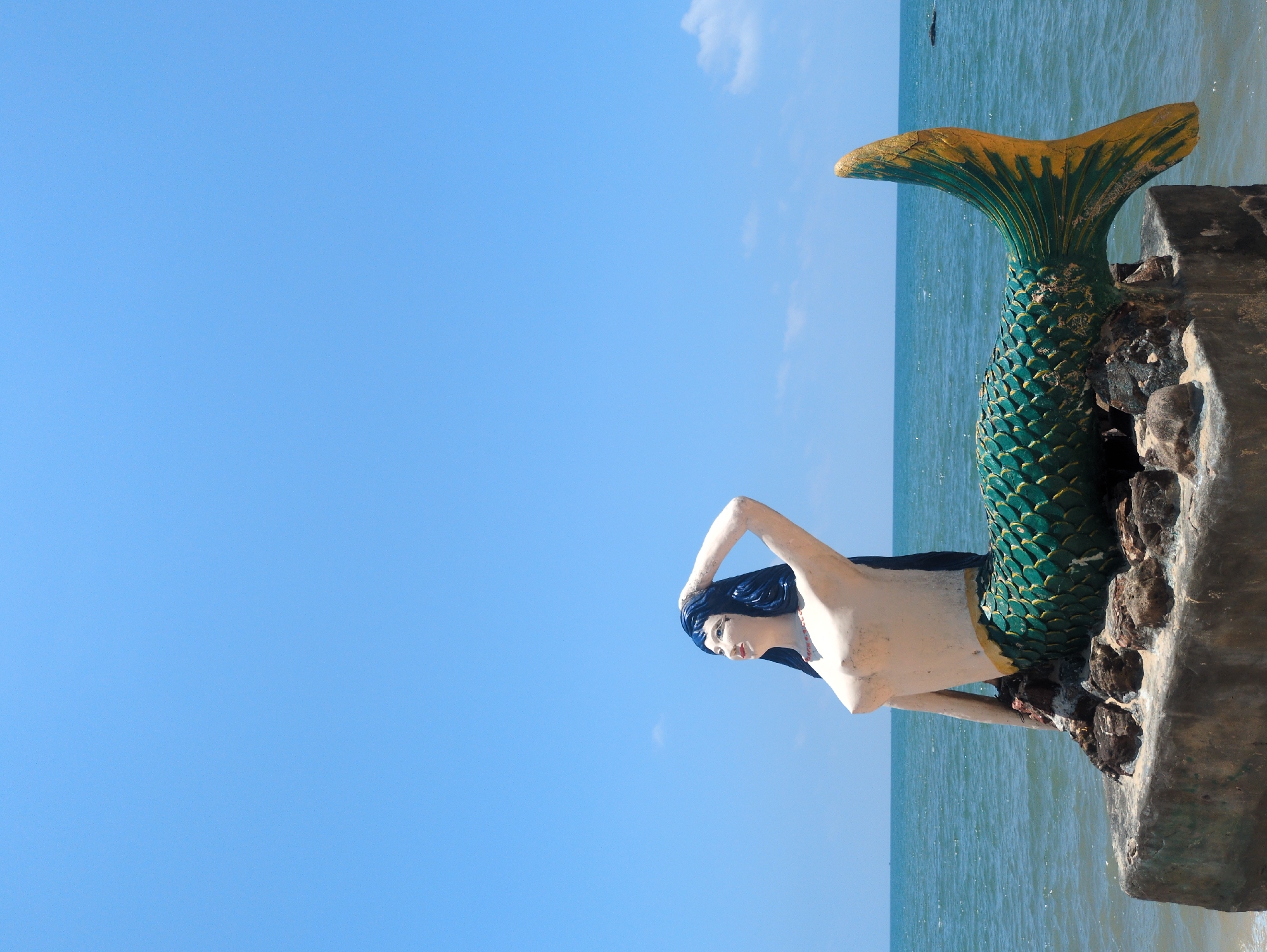 A mermaid statue stands on a concrete block at the edge of the ocean in Phú Quốc, Vietnam. The statue is depicted with a green and gold fish tail. The head of the mermaid is dark blue. The sky is clear, but the image is rotated to the left by ninety degrees.
