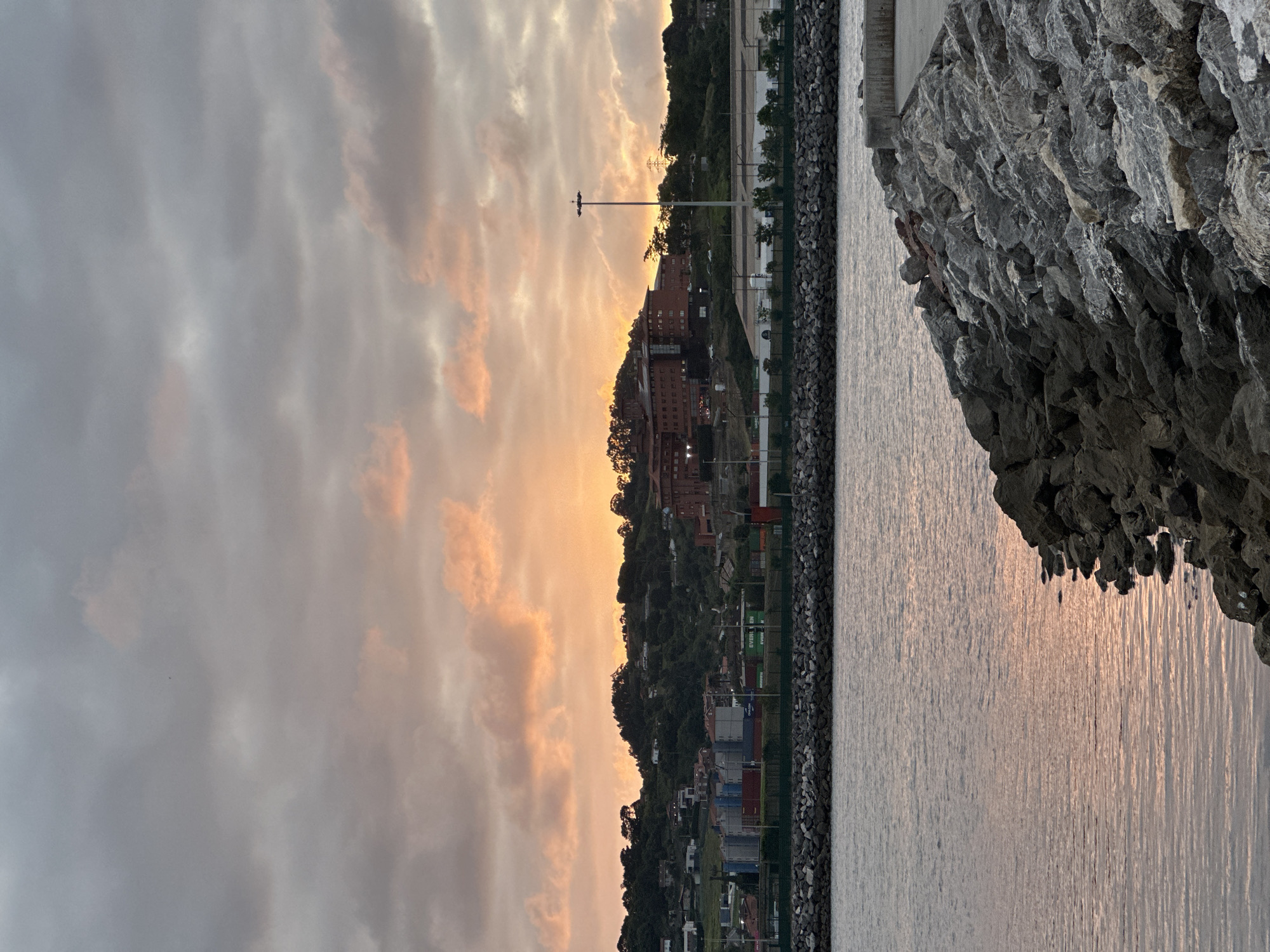 The image depicts a waterfront scene at sunset in Gijón, Spain. The sky is filled with dramatic clouds tinged with orange and pink hues from the setting sun. In the middle ground, a hilly landscape is visible, dotted with buildings and trees. A large, reddish-brown building complex stands out on the hill. Along the coastline, industrial elements such as shipping containers and large buildings are present. The foreground is dominated by a breakwater made of large rocks, with the calm waters of the sea reflecting the colors of the sky. There are no people or specific actions depicted, creating a serene and naturalistic portrayal of the port at dusk.