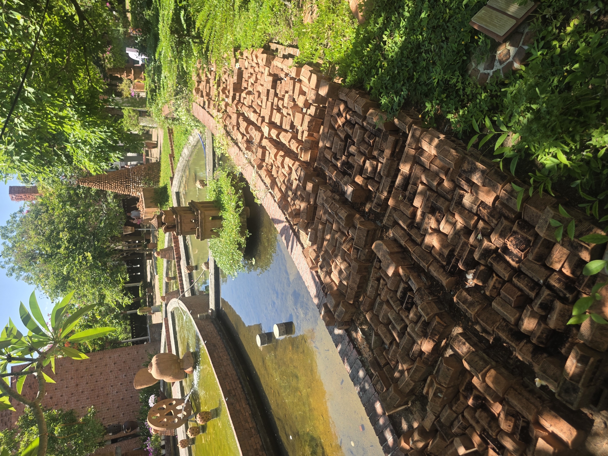 This image presents a vibrant and artfully designed garden or park in Đà Nẵng, Vietnam, bathed in bright sunlight. The scene is characterized by a central water feature, appearing as a meandering canal or pond, bordered by a distinctive wall crafted from numerous reddish-brown bricks. These bricks are uniquely stacked and arranged, creating a textured, sculptural pathway or barrier that runs along the water's edge.
Lush green vegetation frames the scene, including large trees with bright leaves, dense ground cover, and ferns, contributing to a verdant and natural atmosphere. In the foreground, a wider, curved water basin is visible, featuring terracotta or clay decorative elements, including an ornate pot and a circular, wheel-like structure from which water is actively flowing, suggesting a fountain. Further along the main canal, two cylindrical objects are partially submerged in the clear water.
In the background, beyond the water channel, more garden elements are discernible. A tall, narrow structure, also constructed from similar bricks, stands prominently, resembling a decorative tower or chimney. A few indistinct figures, likely visitors, can be seen near this structure, indicating that people are exploring the area.
The bright sun casts distinct shadows, suggesting a clear, sunny day, probably mid-morning or afternoon. A small, light-colored sign or plaque is visible amidst the foliage in the upper right, but any text on it is illegible. The overall impression is one of tranquility, natural beauty, and intricate craftsmanship within a meticulously maintained outdoor setting.