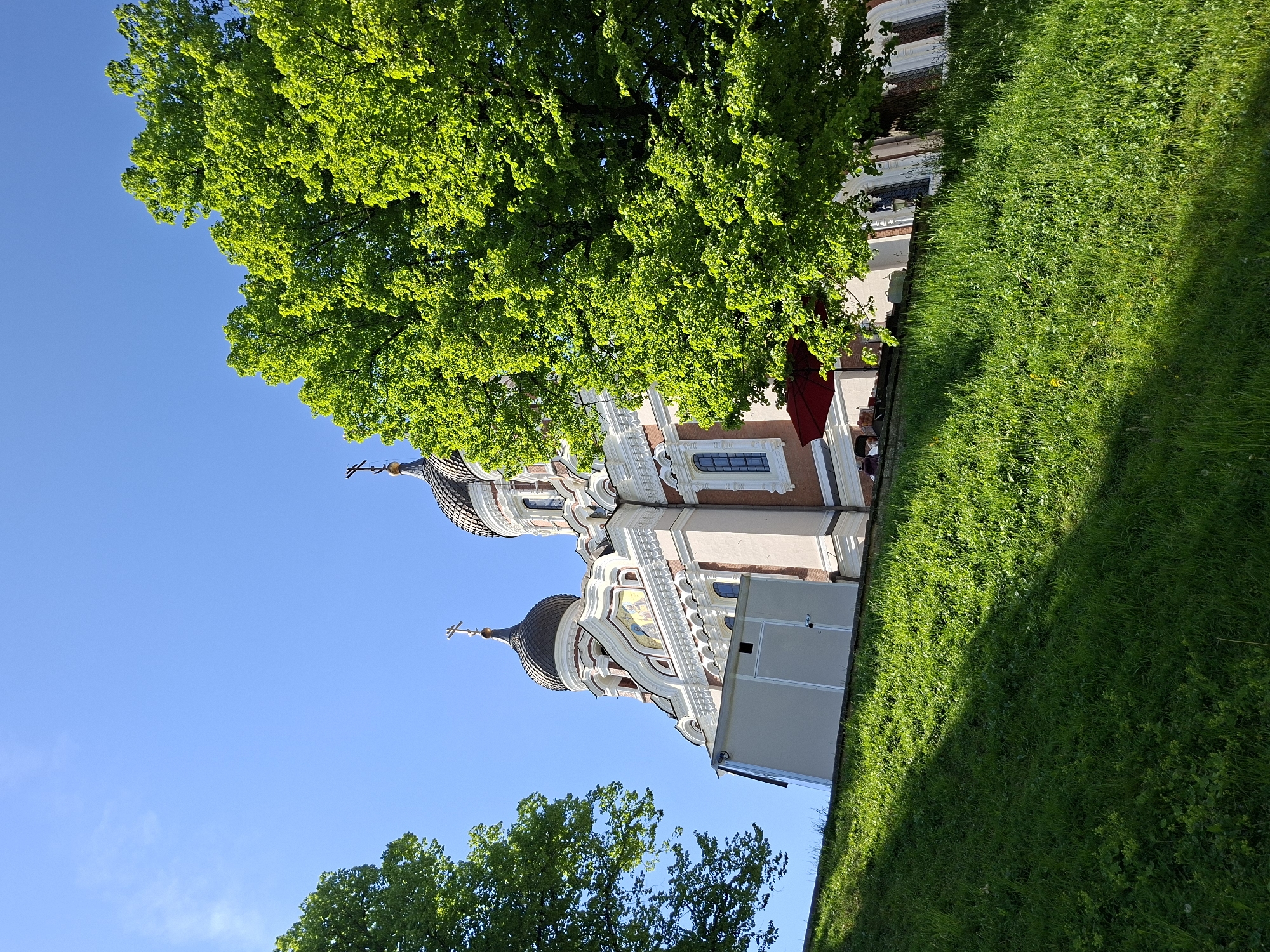 The image captures a vibrant, sunlit view of the Alexander Nevsky Cathedral in Tallinn, Estonia, on a clear day. The architectural masterpiece, characteristic of Orthodox church design, is positioned prominently on the left and central-lower portion of the frame, viewed from a slightly low angle.
The cathedral showcases multiple onion domes, each capped with an Orthodox cross. Its facade is a rich blend of light-colored stone, reddish-brown sections, and intricate white decorative trim, featuring arches, cornices, and decorative patterns. A colorful mosaic or fresco is visible on one of the upper sections of the building. The windows are dark and arched, contributing to the historical aesthetic.
A large tree with lush, bright green foliage dominates the upper left side of the image, extending downwards to partially obscure the cathedral. This vibrant greenery suggests a spring or summer setting. Below this, another smaller section of green leaves is visible. The sky above is a clear, deep blue, indicating excellent weather with minimal cloud cover.
To the right of the cathedral, a manicured slope of bright green grass fills the lower and mid-right sections of the frame. Strong, defined shadows are cast across the grass, pointing to bright sunlight, likely mid-morning or afternoon. There are no people visible, and no specific activities are taking place, presenting a serene architectural and natural landscape. No discernible text is visible within the image.