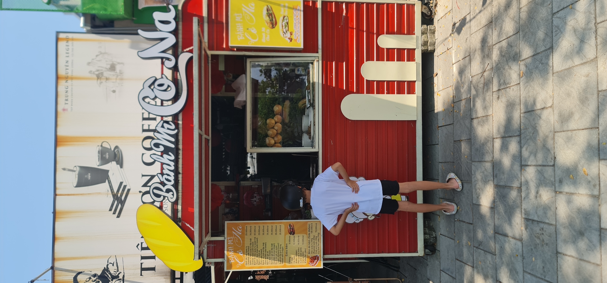 The image shows a street scene in Đà Nẵng, Vietnam, featuring a brightly colored food stall. The stall is primarily red with white accents, prominently displaying the name "Bánh Mì Co Na" in stylized lettering. The stall also has a menu board listing various bánh mì options and prices in Vietnamese Dong. A yellow sign advertises "Bánh Mì Co Na" with a picture of a sandwich.
A person wearing a white shirt, black shorts, a helmet, and flip-flops is standing in front of the stall. The stall's exterior includes depictions of coffee items, suggesting coffee might also be offered. The ground is paved with grey rectangular stones. The sky is visible at the top left corner.