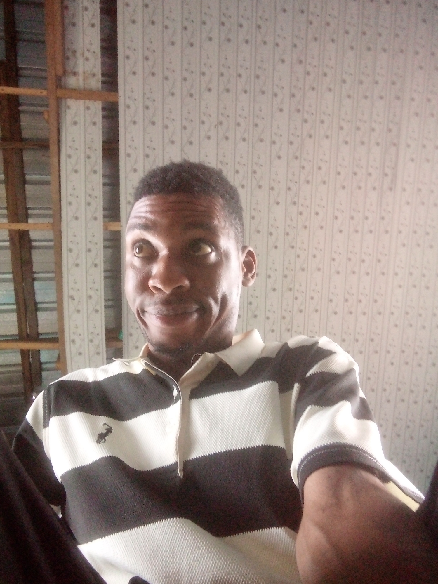 A medium shot captures a dark-skinned man from the chest up. He is wearing a striped polo shirt with black and white horizontal bands and a small logo on the left chest. The background consists of a vertically patterned off-white wall with subtle floral designs and exposed wooden structural elements. The man is looking slightly upwards with a subtle, amused expression on his face. The location is in Ikorodu, Nigeria.