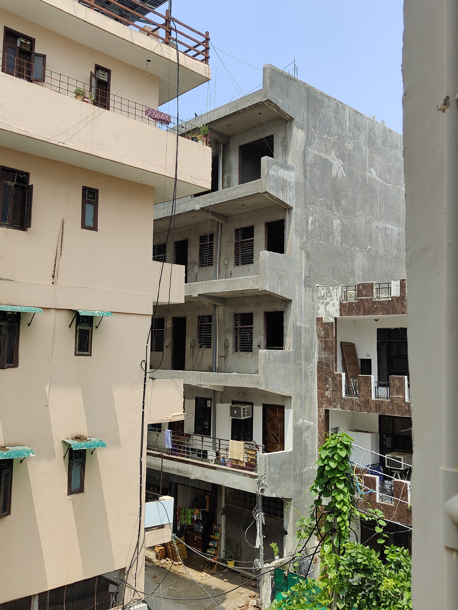 The image captures a street view in Gurgaon, India, characterized by multi-story buildings and construction. On the left, a light-colored building features several balconies with railings and windows. Some windows are open, revealing air conditioning units. Green awnings are visible above some windows on the lower floors. In the center and right, a building under construction exhibits exposed concrete structures and incomplete facades. Brown tiled accents are visible on some balconies of this structure. Lush green foliage, including a tall vine-covered pole and smaller trees, is present in the foreground and along the side of the buildings. Electrical wires are strung between the buildings, and various objects like laundry, potted plants, and what appear to be building materials are scattered around the ground level. The sky is clear and blue, indicating daytime.