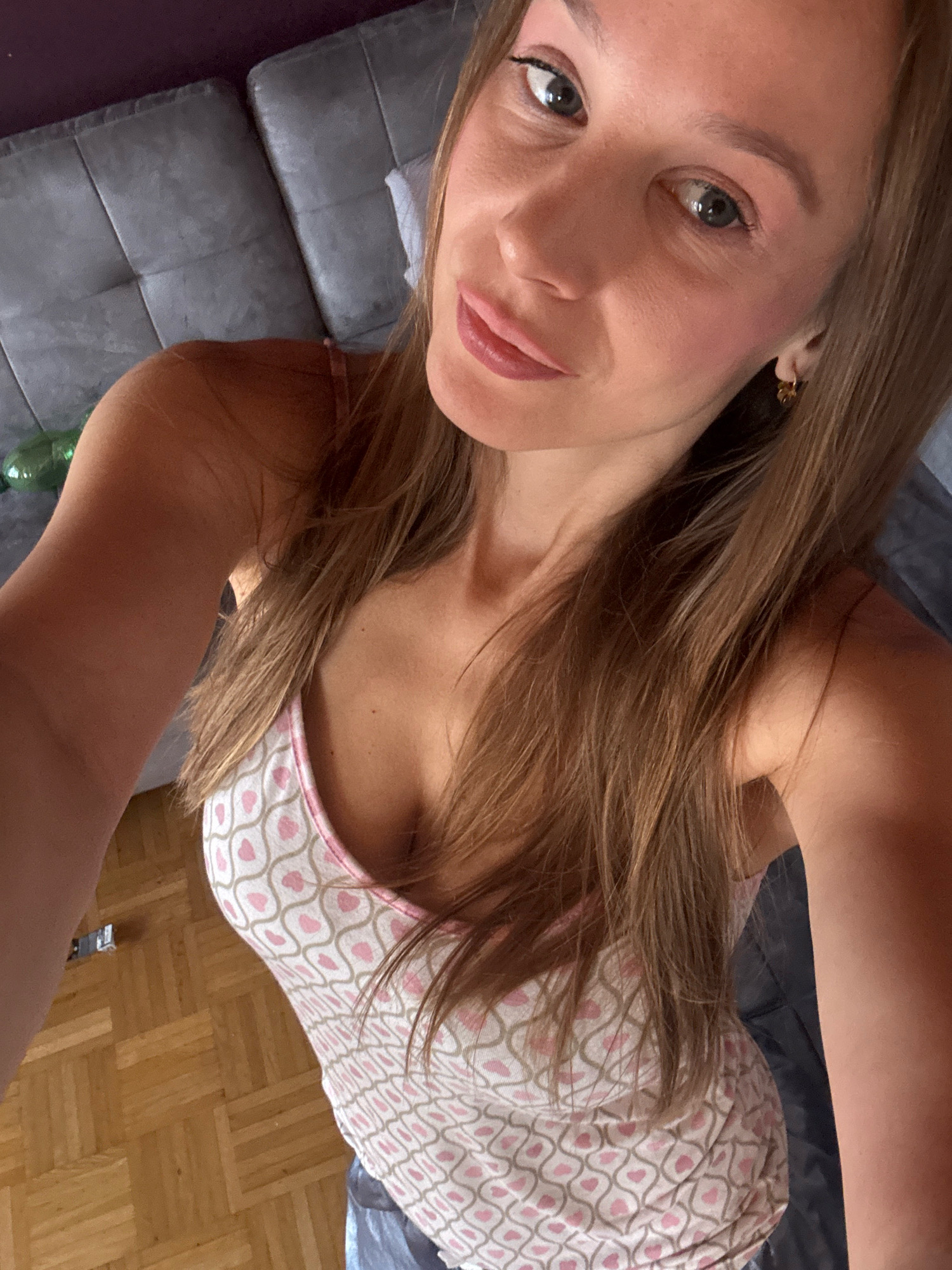 The image shows a young woman indoors, appearing to be in a domestic setting in Überherrn, Germany. She is positioned in the foreground, taking a selfie with her right arm extended. The woman has long, light brown hair that falls around her shoulders. She is wearing a light-colored tank top with a pattern of pink and gold hearts. Her face is angled towards the camera, and she is looking directly into the lens with a neutral expression. Her makeup appears to be natural, with subtle eyeshadow and lipstick.
The background suggests a living area. To the left, a portion of a gray upholstered sofa with a tufted design is visible. In the mid-ground, light brown hardwood flooring with a parquet pattern is present. On the floor, a small silver object, possibly a remote control or electronic device, is located near the edge of the frame. The overall lighting is soft, indicating an indoor environment. The environment appears to be a bedroom or living room.