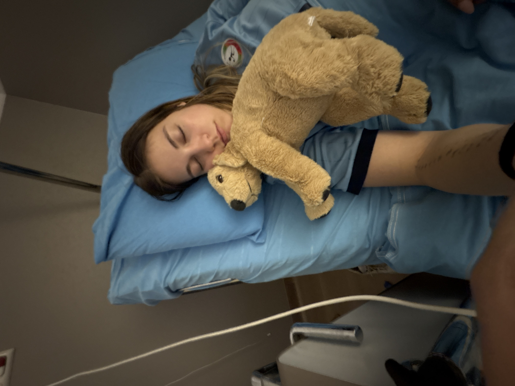 A woman is sleeping on a blue hospital bed with a teddy bear resting on her chest. She has medium-length brown hair and is wearing a blue hospital gown. The bed has blue sheets and a pillow. A medical device and cord are visible near the bed. The location cannot be determined from the image.