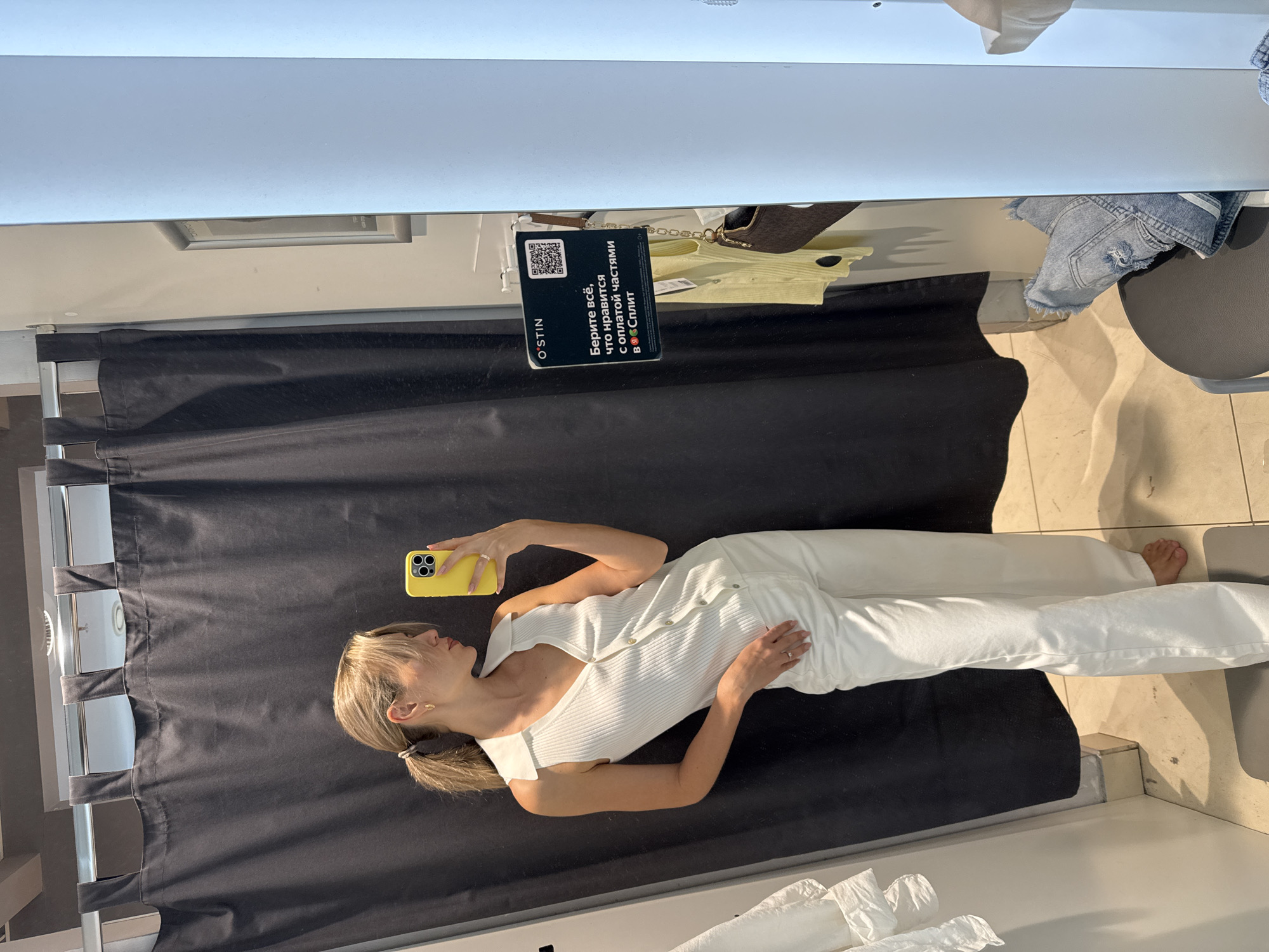 A woman with blond hair is taking a selfie in a clothing store dressing room. She is wearing a white jumpsuit. She is holding a yellow smartphone. The dressing room has a black curtain. A small sign hanging above contains the text "O'STIN" and other text in Cyrillic. There is clothing and a chair visible above the curtain.