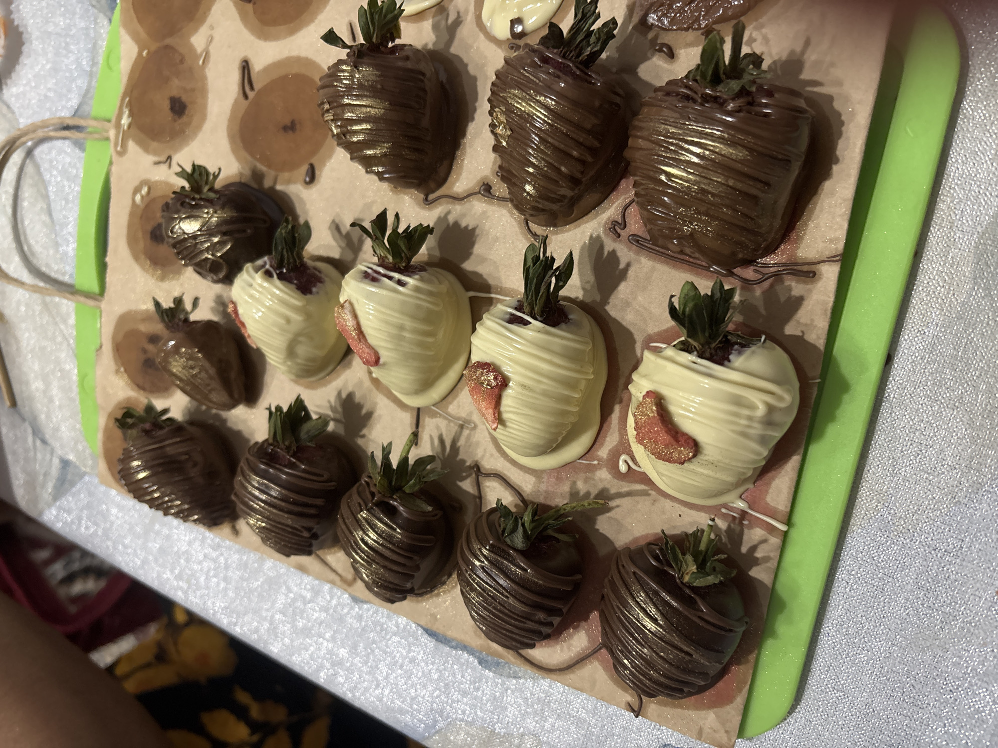 The image displays a collection of chocolate-covered strawberries arranged on a piece of parchment paper, which is laid on top of a green cutting board. Some strawberries are dipped in dark chocolate and drizzled with lighter colored chocolate, while others are dipped in white chocolate and topped with what appears to be dried strawberry pieces and gold edible glitter. A few of the dark chocolate-dipped strawberries also have gold glitter. The arrangement suggests a confectionary preparation or display. The background shows a blurry person and a patterned surface. There is no information within the image that places this scene in the city of Taraz city administration, Kazakhstan.