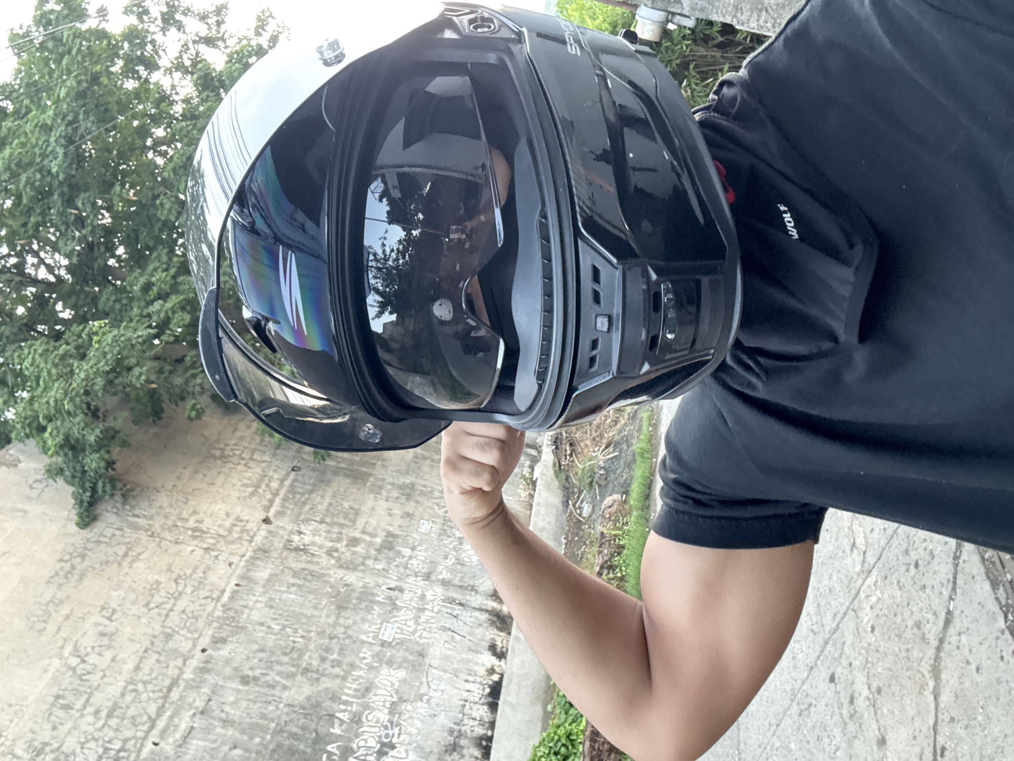 A person wearing a black full-face motorcycle helmet is flexing their bicep. The helmet has a reflective visor showing a partial reflection of buildings and trees. The person is wearing a black t-shirt and a black neck gaiter with the word "WOLF" printed on it. Behind the person, there is a textured concrete wall with some graffiti, and lush green foliage. The ground appears to be a mixture of pavement and dirt. The scene is illuminated by daylight, suggesting it is outdoors. The presence of graffiti on the wall and the general urban outdoor setting are consistent with a location in Davao City, Philippines.