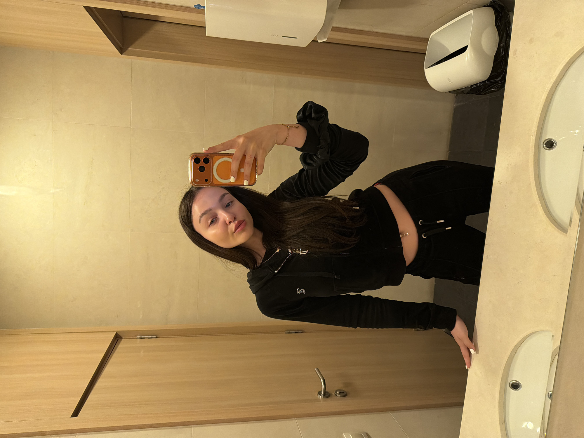 A young woman is taking a selfie in a bathroom. She is holding a smartphone with an orange case. She's wearing a black velvet tracksuit, her hair is long and dark. The bathroom has light beige tiled walls and a sink counter with two round sinks. There is a white hand dryer mounted on the wall and a trash bin next to it. The location is likely a public restroom in Sofia, Bulgaria.