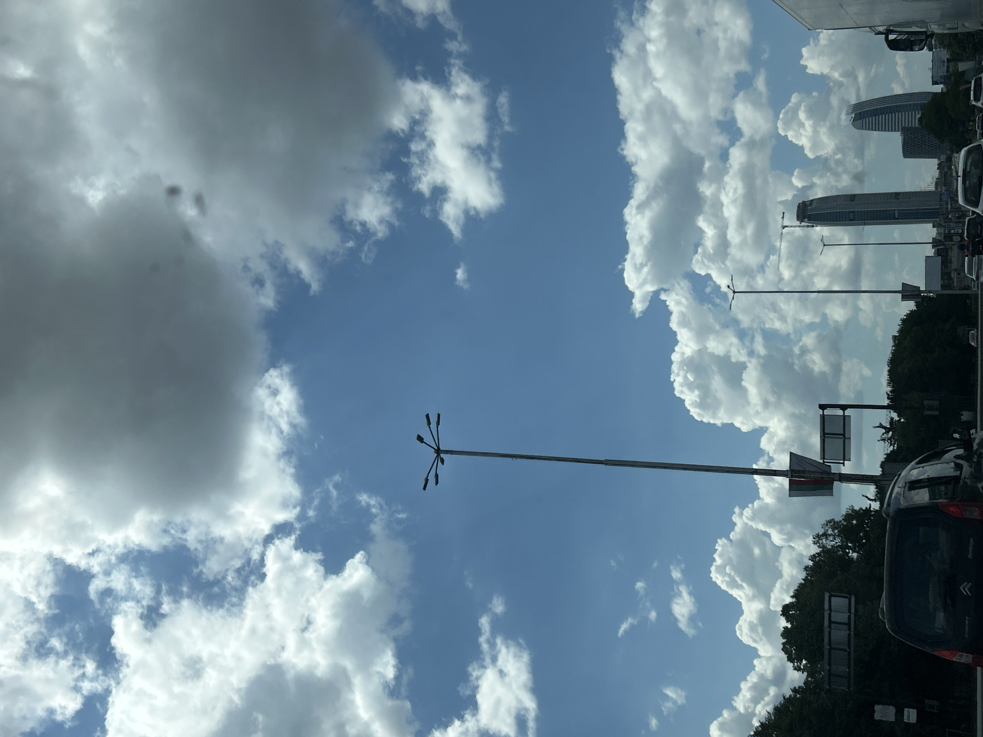The image depicts a bright, sunny daytime scene on a multi-lane road in Sofia, Bulgaria, characterized by a vibrant blue sky dotted with numerous fluffy white cumulus clouds and some larger, darker grey clouds overhead. In the foreground, the rear of a dark-colored Citroën Berlingo vehicle is visible, partially obscuring the lower left portion of the frame.
A tall, grey metallic streetlight pole rises centrally in the midground, featuring a star-like array of six horizontal lamp fixtures at its apex. Further down the road, additional streetlights and a series of rectangular traffic signs are discernible. To the left, a dense line of green trees forms a natural barrier. Notably, in one of the provided hint crops, a vertically hanging Bulgarian flag (white, green, red stripes) is visible attached to a pole, confirming the country. On the far right, part of a large white truck is seen.
The background is dominated by two modern, prominent skyscrapers. The taller, more slender building on the left (identified as Capital Fort) appears to have a construction crane positioned at its summit. To its right, a second, slightly shorter, and distinctively curved skyscraper (identified as Sky Fort) stands. The scene includes other vehicles on the road in the distance.
Based on the distinctive architecture of the Capital Fort and Sky Fort skyscrapers, the location is identified as the area around Business Park Sofia, specifically visible from Tsarigradsko Shose boulevard, looking generally towards the east/southeast near the intersection with the Sofia Ring Road (Okolovrasten pat).