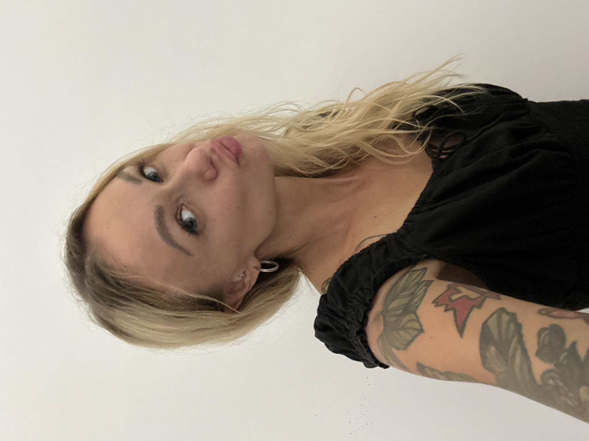 A woman with blonde hair and blue eyes is posing in a black top. She has a tattoo on her left arm. The background is a plain white wall. This location information cannot be determined from the image.