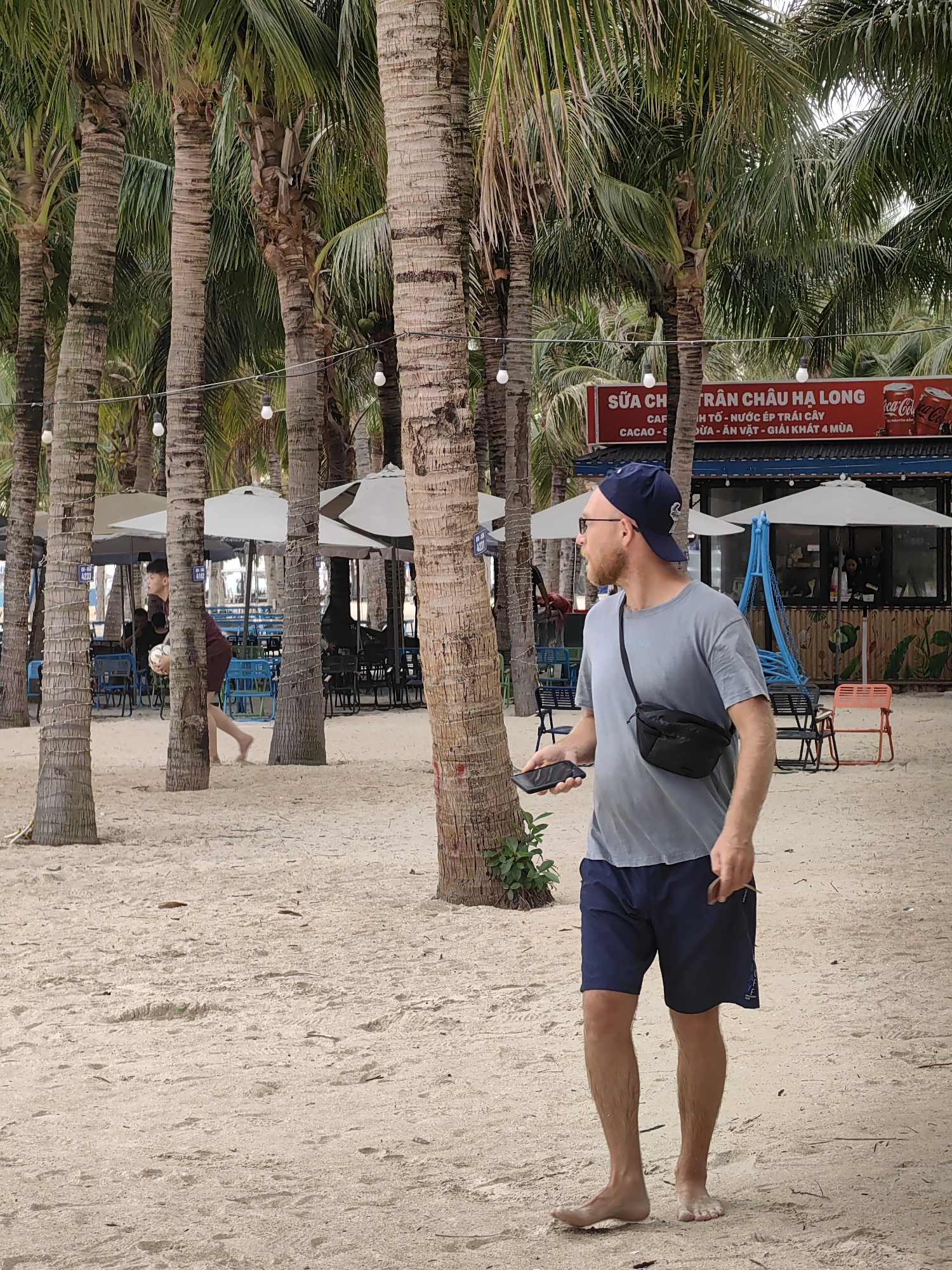 The image depicts a casual daytime scene at a sandy beachside area in Bai Chay Ward, Vietnam. Numerous tall palm trees with string lights strung between them line the sandy ground.
The primary subject is a fair-skinned man with a beard, wearing a blue baseball cap, a grey t-shirt, dark blue shorts, and a black fanny pack. He is barefoot and walking on the sand, holding a black smartphone in his left hand, and looking to his left, seemingly observing his surroundings. In the mid-ground, another person, a young male, is visible walking between palm trees, carrying a white ball.
In the background, there are white beach umbrellas, blue and orange seating arrangements, and a red and blue refreshment kiosk. The kiosk has signage in Vietnamese, which reads "SỮA CHUA TRÂN CHÂU HẠ LONG" (Ha Long Pearl Yogurt/Milk Tea), "CÀ PHÊ" (Coffee), "CACAO", "NƯỚC ÉP TRÁI CÂY" (Fruit Juice), "DỪA" (Coconut), "ĂN VẶT" (Snacks), and "GIẢI KHÁT 4 MÙA" (Four Seasons Refreshment). Coca-Cola branding is also visible on the kiosk.
The weather appears to be overcast or hazy, creating diffused light. The overall atmosphere suggests a relaxed, leisurely day at a tropical or coastal destination.