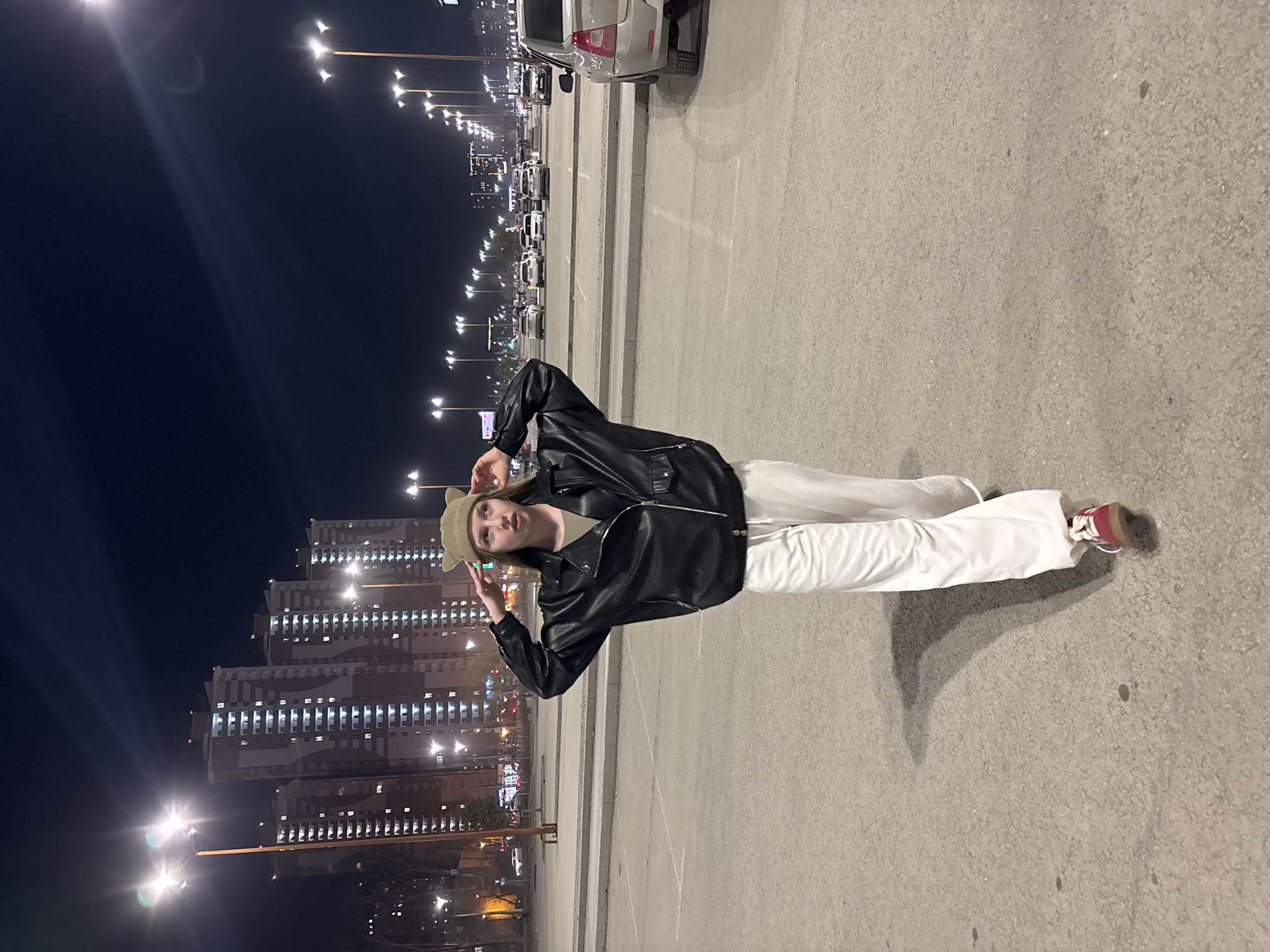 A young woman stands in a parking lot at night in Perm, Russia. She wears a black leather jacket, white pants, and a tan knit cap. She is making a hand gesture near her head. To her left are several tall apartment buildings with illuminated windows. Behind her is a row of parking spaces, some occupied by vehicles. Streetlights line the area and cast light on the scene. The parking lot surface is concrete with painted parking lines.