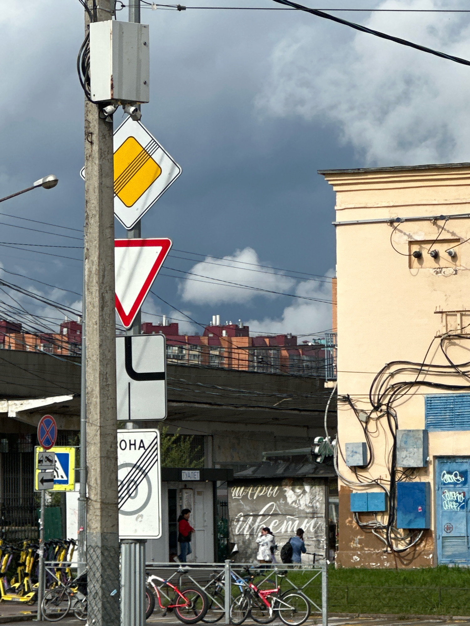 The image depicts a street scene in Murino, Russia, under a cloudy sky. A concrete utility pole dominates the foreground, adorned with various traffic signs. The uppermost sign is a yellow diamond with a thick black border and three horizontal black lines. Below it is a red and white inverted triangle, a yield sign. Further down the pole is a white rectangular sign with a black L-shaped arrow indicating a turn. Another rectangular sign to the right of the pole shows the Russian text "ОНА" (ONA), likely indicating a pedestrian crossing or a specific zone, with a circular sign superimposed with diagonal lines, possibly a speed limit or no entry indicator. Adjacent to this is a blue and yellow diamond-shaped sign with a black arrow pointing left and a black horizontal bar, suggesting a lane restriction.
To the right of the pole is a light beige building with a dark grey roofline. The building's facade is covered in a dense network of black electrical wires and conduits, connecting to several blue electrical boxes mounted on the wall. A blue metal door with graffiti is visible on the right side of the building. In front of the building, a small kiosk-like structure has signage with white cursive lettering.
In the middle ground, an elevated road or overpass is visible, with buildings of red and orange brick construction in the background. On the left side of the image, a row of yellow electric scooters is parked. In front of the railing in the foreground, several bicycles are parked, including one with a red frame and pink rims.
Three people are present in the scene: a woman in a red jacket and jeans is standing near the kiosk. Two individuals, one in a white jacket and the other in a denim jacket and backpack, are conversing near the entrance of the kiosk. The overall environment suggests an urban setting with a mix of infrastructure and pedestrian activity.
