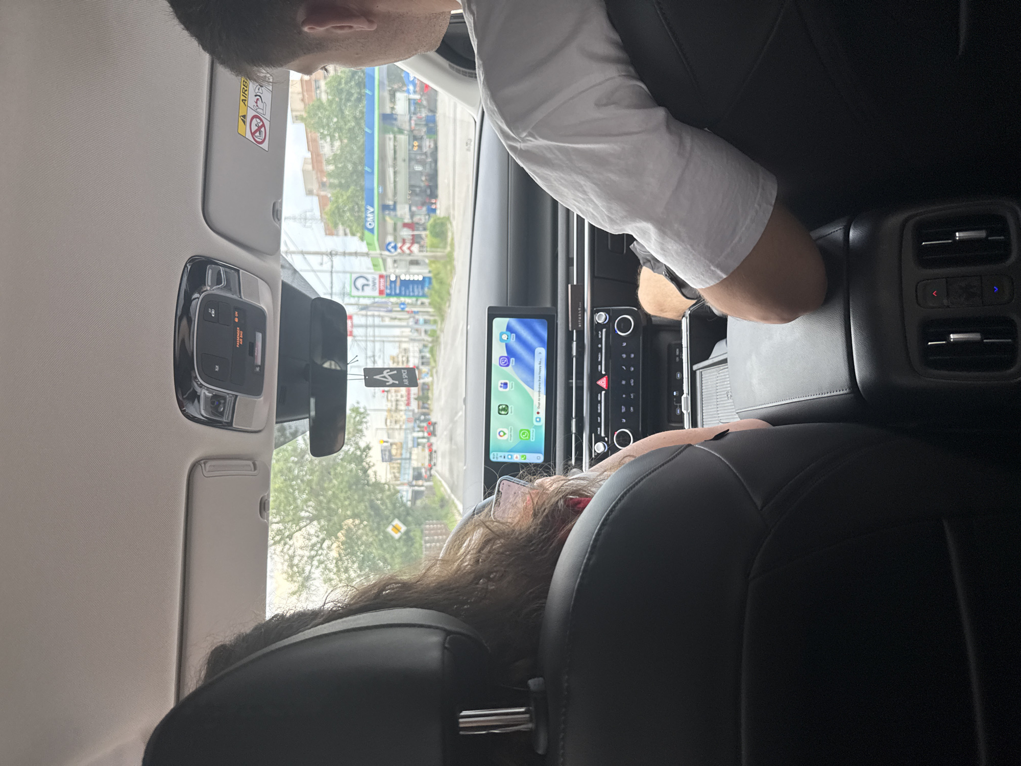 The image, captured from the rear passenger seat of a vehicle, displays the front cabin and the view through the windshield onto an urban street in Sofia, Bulgaria, specifically at the OMV petrol station located at Boulevard "Tsar Boris III" 215. In the left foreground, the back of a front passenger's head is visible, characterized by long, dark, wavy hair. This individual holds a smartphone in their hand, resting on their lap, displaying a home screen with various application icons, including WhatsApp, Maps, and Instagram, along with "Gmail." A pink-tinted object is visible near their head. In the right foreground, the right side of the driver's head and shoulder are visible; the driver wears a white short-sleeved t-shirt, and his right arm rests on the center console.
The car's interior features black leather seats and a dashboard equipped with a wide central infotainment screen, which displays a similar array of app icons and a partial message reading "Drear Air conditioning from Happy Be...". Below the screen are digital climate controls and physical buttons. A "RITUALS..." branded air freshener or diffuser is mounted on the dashboard. An "AIR SPACE" air freshener hangs from the rearview mirror. The overhead console contains interior lighting controls and a "PASSENGER AIR BAG ON" indicator. The right sun visor displays an "AIRBAG" warning label. In the rear cabin, air vents and temperature controls (red for hot, blue for cold) are visible on the center console.
Through the windshield, an overcast sky hangs over a multi-lane road. On the right, a prominent OMV petrol station with its characteristic blue and green canopy and branding is clearly visible, positioned in front of a multi-story residential building. On the left side of the road, further down, street signs including a yellow diamond-shaped priority road sign are present. Other vehicles are visible in the distance on the road.