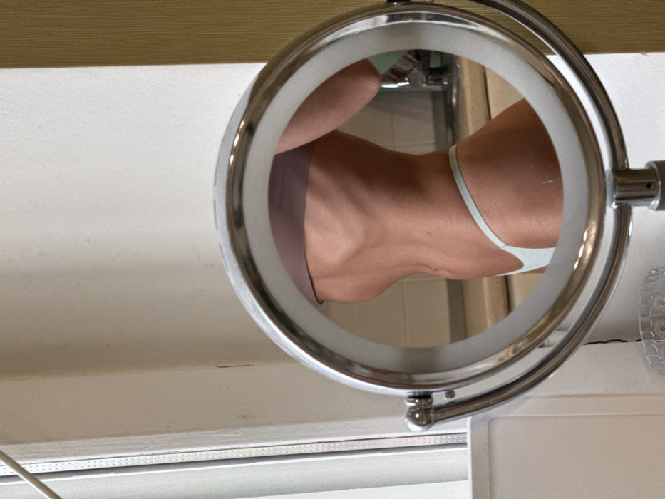 The image displays a close-up indoor scene, centered on the reflection of a person's midsection and upper thighs in a round, illuminated vanity mirror with a chrome frame. The individual is shown in profile, facing right, wearing a light purple bandeau top and light blue thong-style underwear. Their skin appears tanned, and the torso exhibits visible rib and abdominal musculature. The mirror's outer rim is lit, suggesting an LED light ring.
Behind the mirrored reflection, an indoor setting is visible, featuring a sink faucet and a light-colored countertop, possibly indicating a kitchen or bathroom. Surrounding the mirror stand, the foreground shows a white wall and a white window frame to the left, and a textured, golden-beige wall to the right. A clear, faceted glass object is partially visible below the mirror stand. The specific location within the city of Saint Petersburg, Russia, cannot be determined from the visual evidence provided.