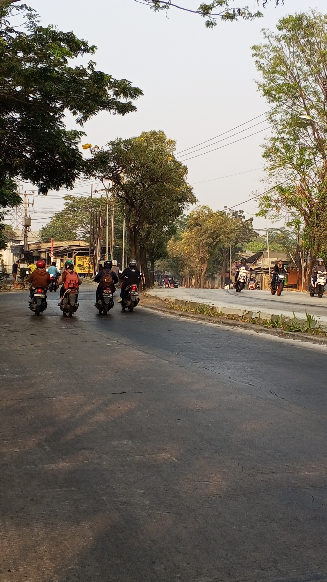 The image depicts a street scene in Bekasi, Indonesia, with several motorcycles and their riders moving along a paved road. The road is bordered by trees and vegetation on both sides, and some buildings are visible in the background. The sky is clear and bright, suggesting it is daytime.
In the foreground, four motorcycles are traveling away from the camera, with riders wearing helmets and casual clothing. One rider in front has a red backpack, and the rider behind them has an orange backpack. Further down the road, more motorcycles are visible, some in groups, heading in the same direction. One motorcycle in the distance has a green body.
The environment appears to be a semi-urban or suburban area, with a mix of residential buildings, utility poles, and natural greenery. The road shows some signs of wear and tear, with cracks and uneven surfaces. The overall impression is one of everyday life in a busy Indonesian city.