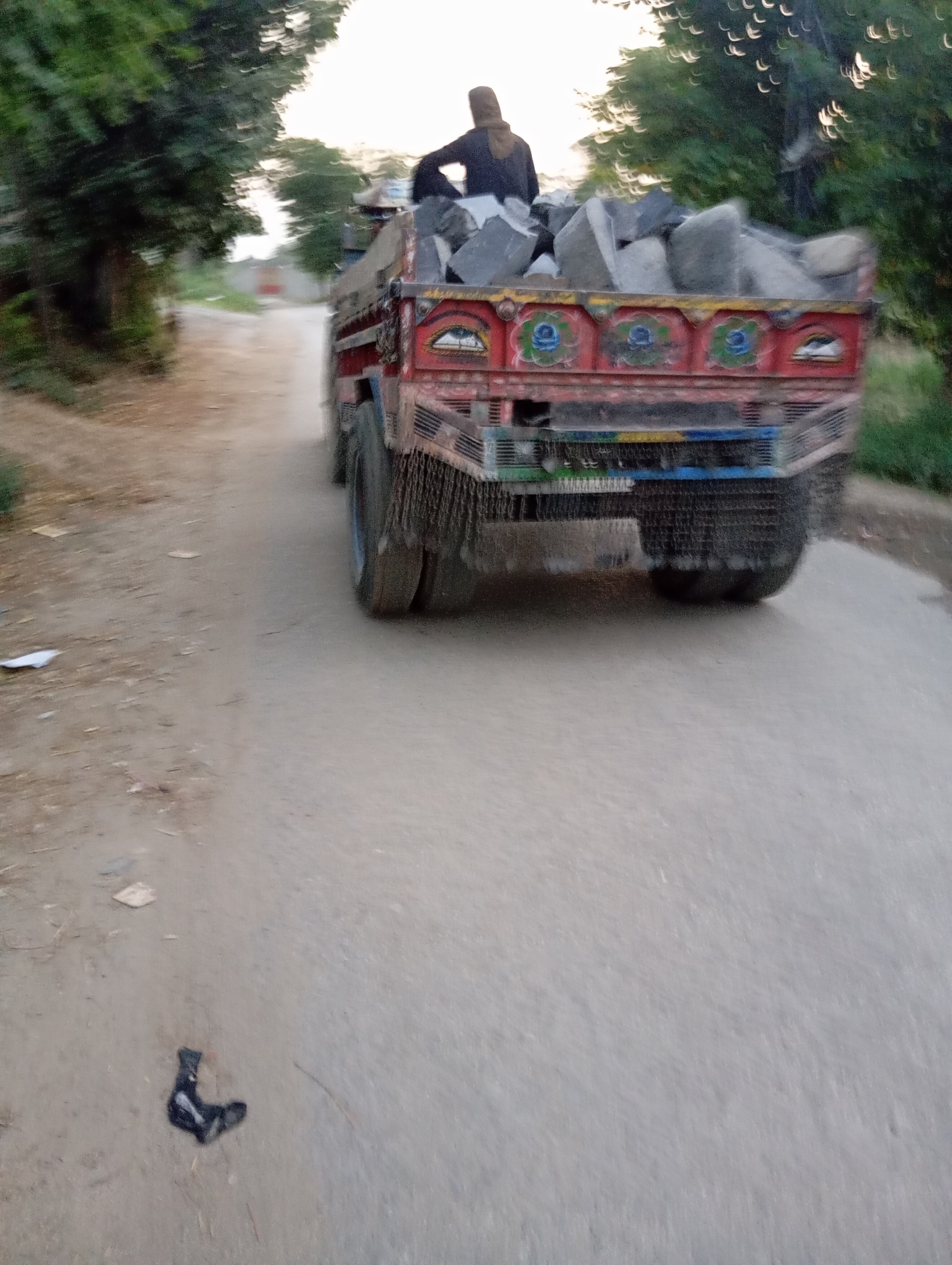 A truck carrying large stones is moving down a dirt road in Lassan, Pakistan. The truck is decorated with colorful floral patterns. A person wearing a brown scarf and dark clothing is seated on top of the stones. The road is lined with trees and appears to be in a rural or semi-rural area. There is some litter on the side of the road, including a black plastic bag. The image is blurred due to motion.