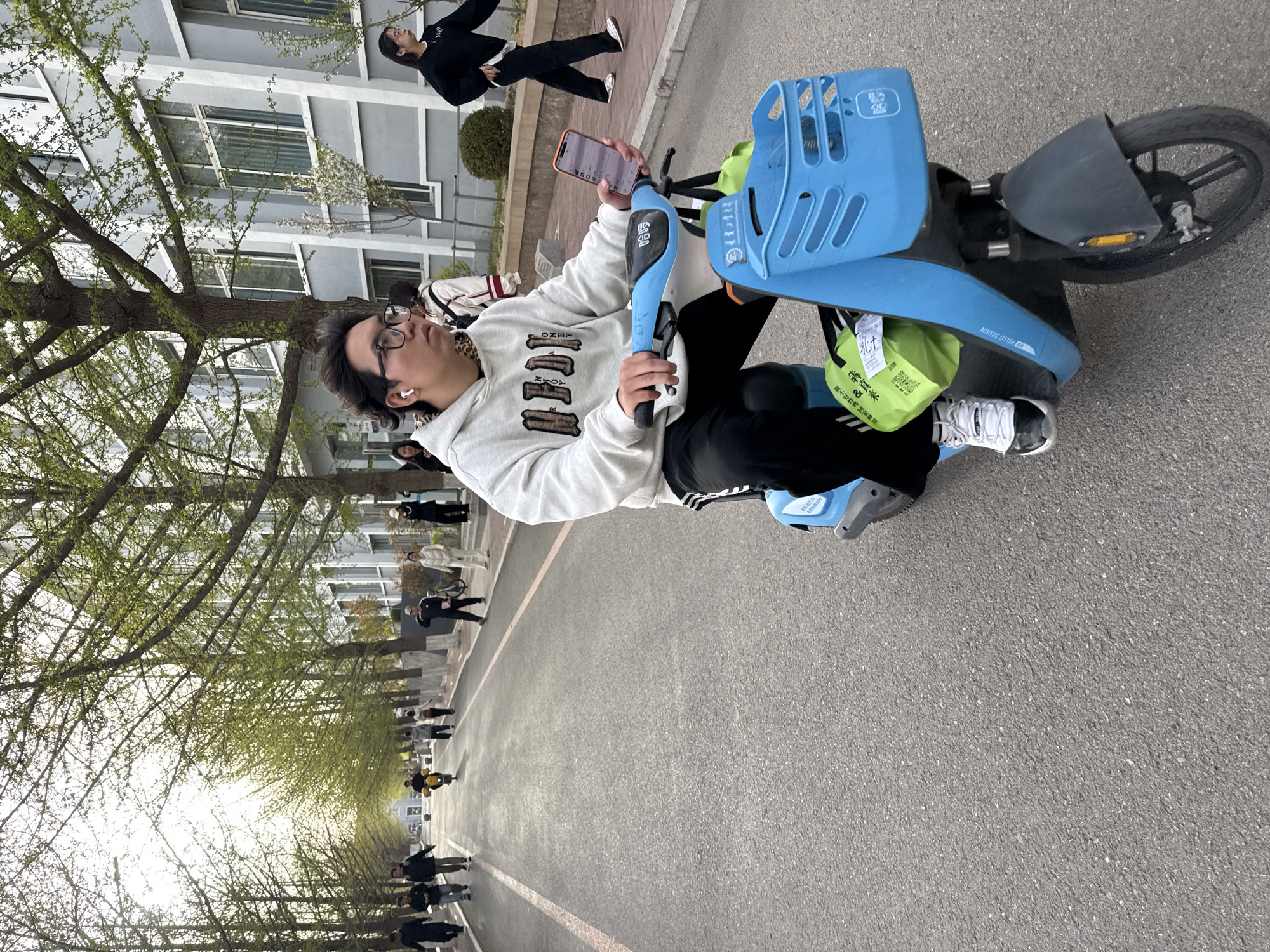 An outdoor, rotated image depicts a young man seated on a blue and black shared electric bicycle in Zhongtun, China, during the daytime. The man, wearing glasses, a white hoodie, black pants, and white sneakers, has white AirPods in his ears and is holding an orange-cased smartphone, looking at its screen. His hoodie has dark block letters that appear to spell "ONEY" and "NJOY," likely part of a phrase.
The shared e-bike features a blue front basket and handlebars. Visible text on the bike includes "哈啰" (Hello) and "HELLO DESIGN." A bright green plastic bag, hanging from the front of the bike, displays Chinese characters such as "犇真来" and "不好炸鸡家神田," possibly advertising or product information.
The setting appears to be a campus or a wide pedestrian street, with a paved road marked by a white line. Rows of trees with fresh, light green leaves line both sides of the path, suggesting it is spring. In the background, several people are walking. Modern, multi-story grey buildings with numerous windows are visible behind the trees and pedestrians. The lighting is bright, indicating it is daytime.