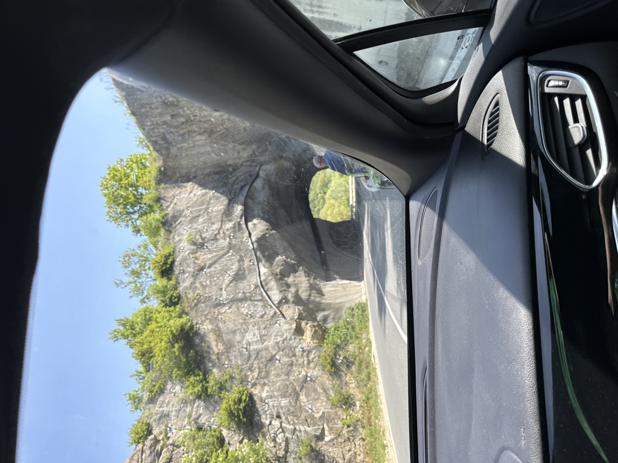 From inside a car, a tunnel entrance is visible on a sunny day. The tunnel is built into a rocky cliff face, which is covered with wire mesh and some vegetation. A road runs alongside the cliff. The car's interior, including the dashboard, air vents, and part of the window frame, is in the foreground. The location is likely near Bilka, Bulgaria, based on the presence of a tunnel through a rocky landscape.