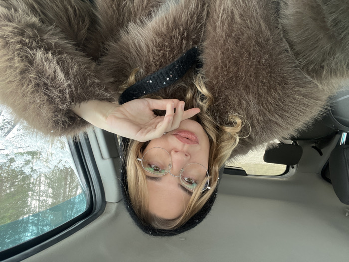 A single female is depicted in a front-facing selfie taken from inside a vehicle in Ust-Ilimsk, Russia. The woman has blonde, wavy hair and wears silver-framed round glasses. Her lips are full and glossy. She is wearing a dark, possibly sequined, beanie and a substantial brown fur coat. Her left hand is raised, with fingers resting against her chin/jawline. The vehicle's interior shows dark headrests in the background. Through the passenger-side window, a snow-covered environment with trees is visible, indicating winter conditions.