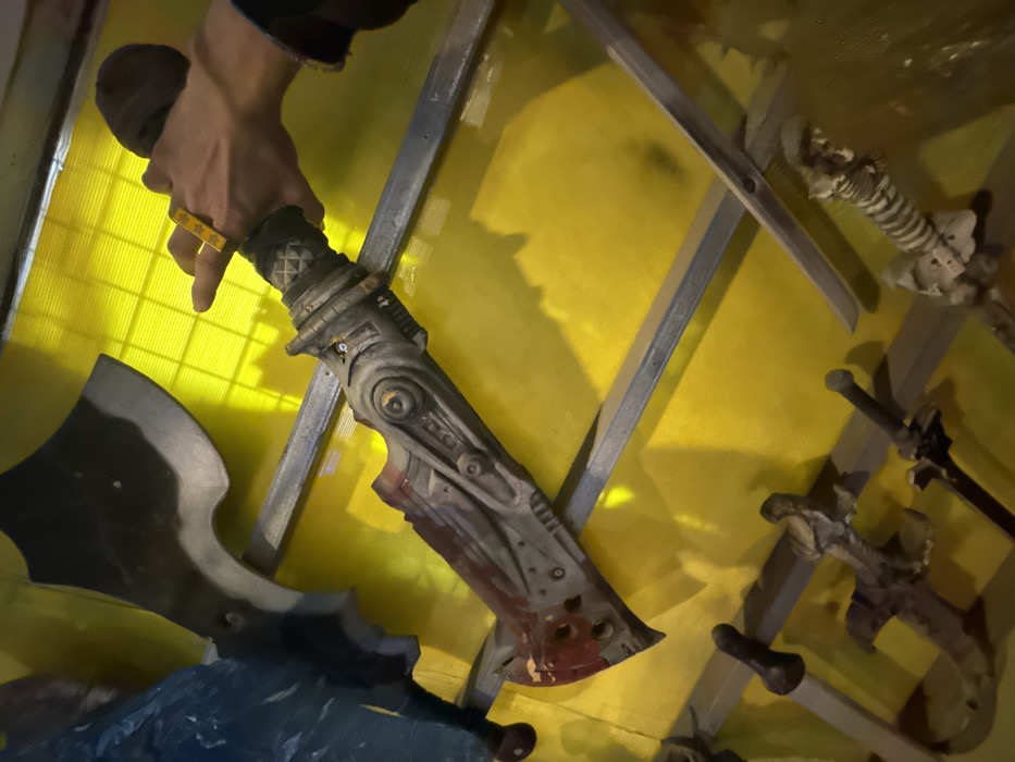 This media file presents an indoor scene located in Tel-Aviv, Israel, showcasing a collection of themed props. The primary focus is a person's right hand and forearm, holding a large, intricately designed weapon. This weapon appears to be a fantasy-style axe-sword or a heavy blade, featuring a robust, mechanical-looking handle with a patterned grip. The blade itself exhibits a reddish, blood-like stain near its edge, contributing to its grim aesthetic. The individual's hand is adorned with a yellow ring featuring small star shapes on the ring finger.
The background consists of a vibrant yellow, possibly corrugated or textured, surface. Against this backdrop, grey metallic or painted shelving units are diagonally mounted, serving as a display for additional props. These include several other bladed weapons resembling swords and daggers. In the upper right corner, a prop designed like a skeletal spine or ribcage is also visible. The overall setting suggests a display within a themed environment, such as a gaming venue, a specialty shop, or an exhibition related to fantasy, horror, or medieval themes. The lighting is artificial, casting shadows across the yellow background. No visible text is present in the image.