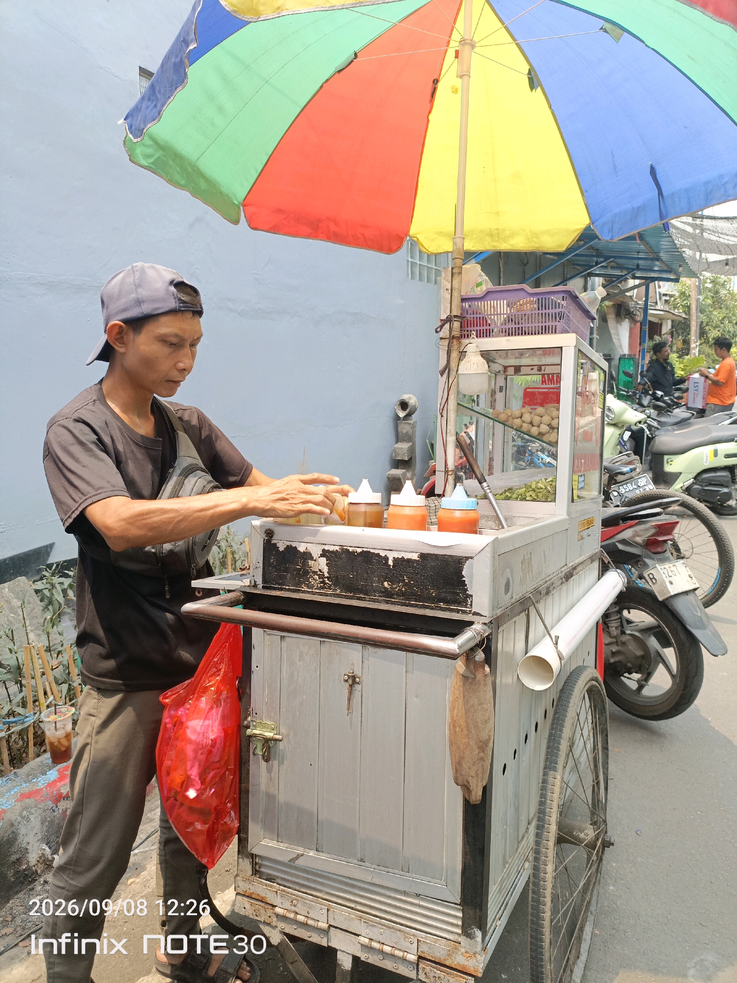 A street vendor is preparing food at a mobile cart in Central Jakarta, Indonesia. The vendor, a man wearing a black t-shirt and a gray baseball cap, is focused on his task. He stands behind a silver metal cart that is equipped with a glass display case containing food items and bottles of condiments. A large, colorful umbrella, divided into sections of red, yellow, blue, and green, provides shade over the cart and vendor. In the background, a few motorcycles are parked, and two other individuals are visible further down the street. The scene appears to be on a paved street with a blue-gray wall behind the vendor and some vegetation to his left. A red plastic bag hangs from the side of the cart. The bottom of the image contains a timestamp: "2026/09/08 12:26" and "Infinix NOTE30".