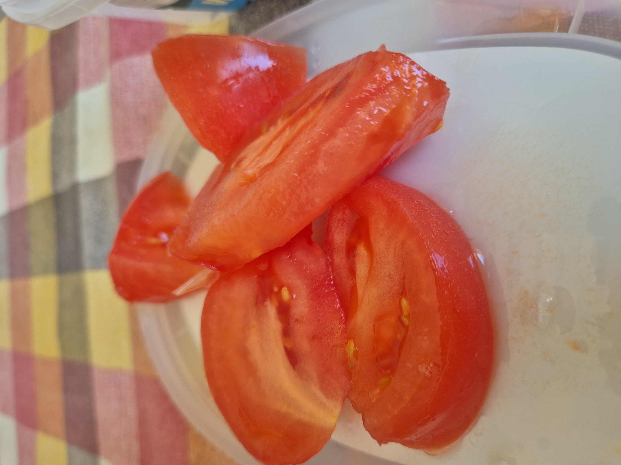 The image shows several slices of a red tomato on a white plate. The tomatoes are cut into wedges, and their interior flesh, seeds, and juice are visible. The plate is on a table covered with a checkered tablecloth featuring alternating bands of red, yellow, and a muted pink or grey. The lighting suggests an indoor setting, possibly a kitchen or dining area. The background is out of focus, but it shows the pattern of the tablecloth extending beyond the plate. There are no people or animals present.