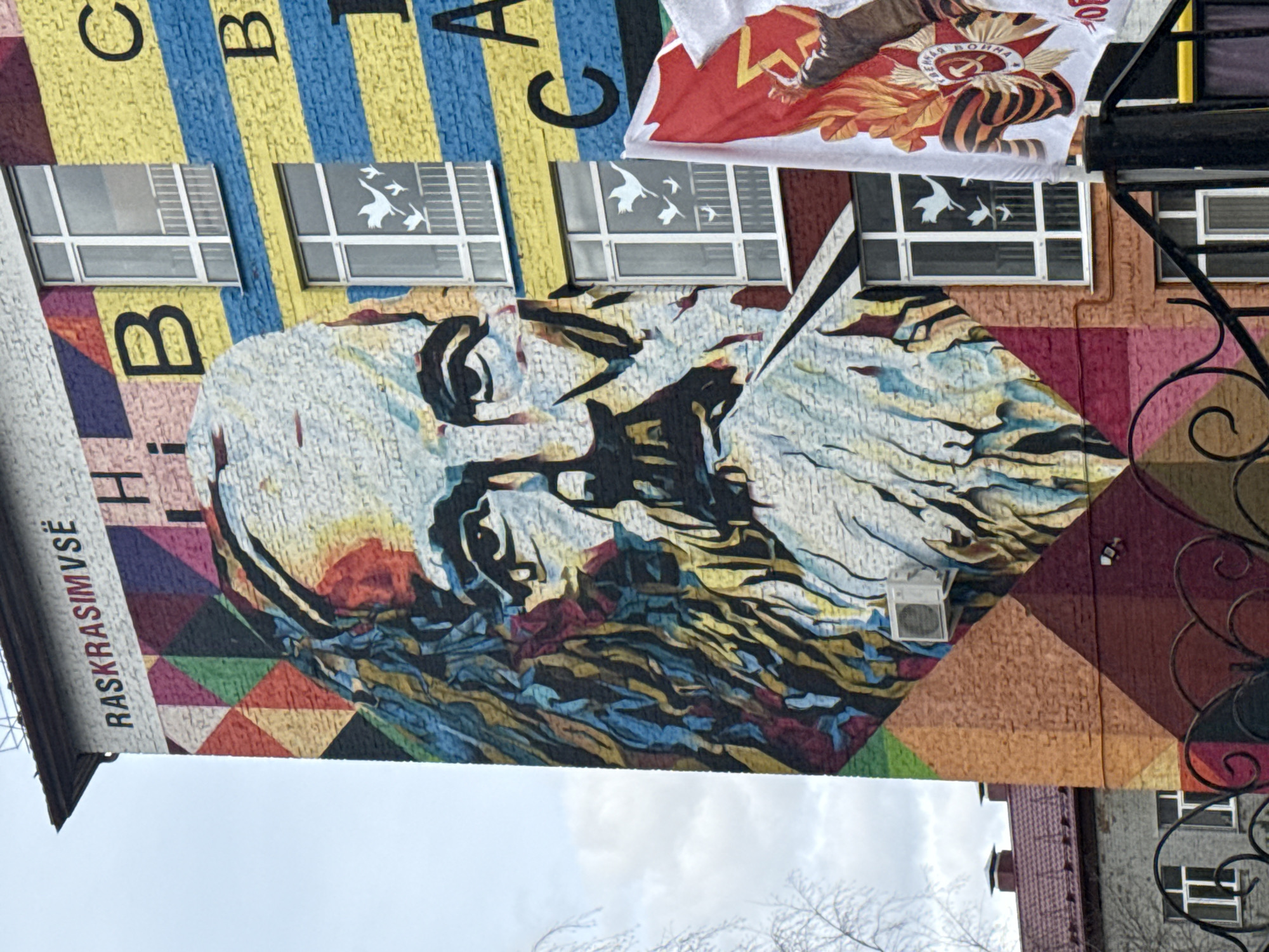 The image shows a mural on a building in Tobolsk, Russia. The mural features a large portrait of a man with a beard, possibly a historical figure, rendered in a textured, mosaic-like style. The portrait is placed on a background of geometric shapes and colors, including triangles in shades of red, orange, yellow, green, and blue. Above the portrait, the building is painted with vertical stripes of blue and yellow, interspersed with white window frames. Some of the windows have stickers showing silhouettes of flying birds. A white banner with red and orange designs hangs near the top of the building on the right side of the frame. The text "RASKRASIMVSË" is vertically written on the left side of the building. Part of another building and wrought iron detail are visible on the right. The sky appears overcast.