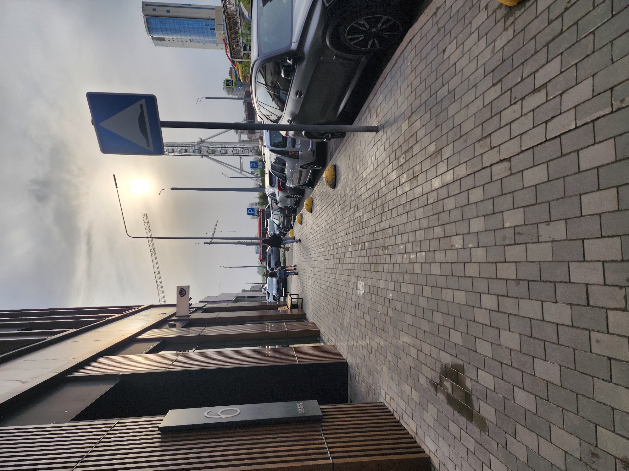 The image shows a street scene in Kazan, Russia, notable for its combination of modern architecture and infrastructural elements. A row of parked cars, mostly light-colored, is aligned along the left side of the street, separated from the pedestrian walkway by yellow bollards. A blue and white road sign, likely indicating an uneven road surface, is positioned near the parked cars. A high-rise building can be seen in the background, suggesting an urban setting. The pedestrian walkway is paved with gray bricks and appears well-maintained. In the distance, there are a few people walking on the sidewalk. A modern building is on the right, with a dark facade and horizontal wooden slats near the ground. A sign with a white circle is visible on the building. The sky is overcast, and the sun is visible but diffused. A crane is visible in the background.