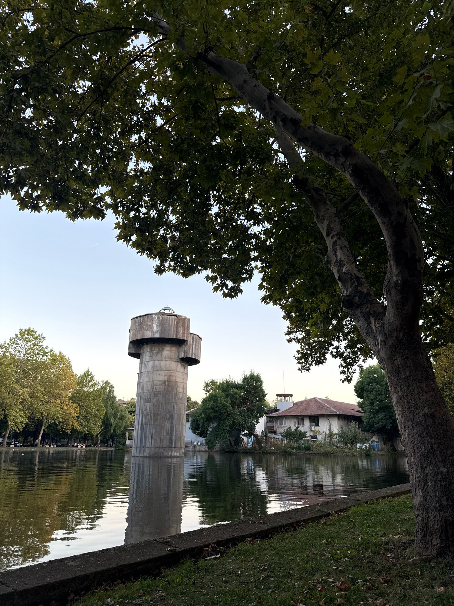 The image depicts a serene waterside scene with a distinctive concrete water tower as the primary subject. The tower is tall and cylindrical, with a rounded top section featuring multiple tiers and a small dome-like structure. It is situated on the edge of a calm body of water, likely a lake or river, whose surface reflects the sky, trees, and the tower itself. The water is bordered by a grassy bank with a concrete edge in the foreground, and further back, a line of trees with lush green foliage. Beyond the trees, a building with a red-tiled roof and white walls is visible, adding to the tranquil, park-like atmosphere. The sky is clear and pale, suggesting it might be late afternoon or early morning. The overall impression is one of quietude and natural beauty, possibly within an urban park setting in Montana, Bulgaria, given the water tower's architectural style which is characteristic of such infrastructure in Eastern Europe.