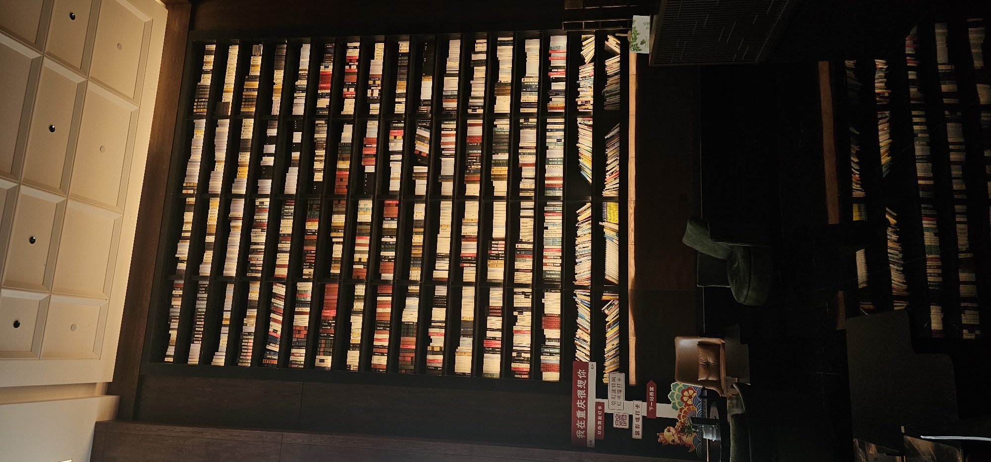 This photograph shows a large wall of books in a library or bookstore. The books are arranged vertically in rows and columns. The shelves are dark and the books appear to be illuminated from behind. In the lower right corner, there is a set of signs written in Chinese characters. The words are "我在重庆很想你" (I miss you in Chongqing), along with promotional information for the bookstore. The setting is likely located in the city of Liangjiang, China. There are chairs and couches in the background suggesting a lounge area. The wall to the left is white.