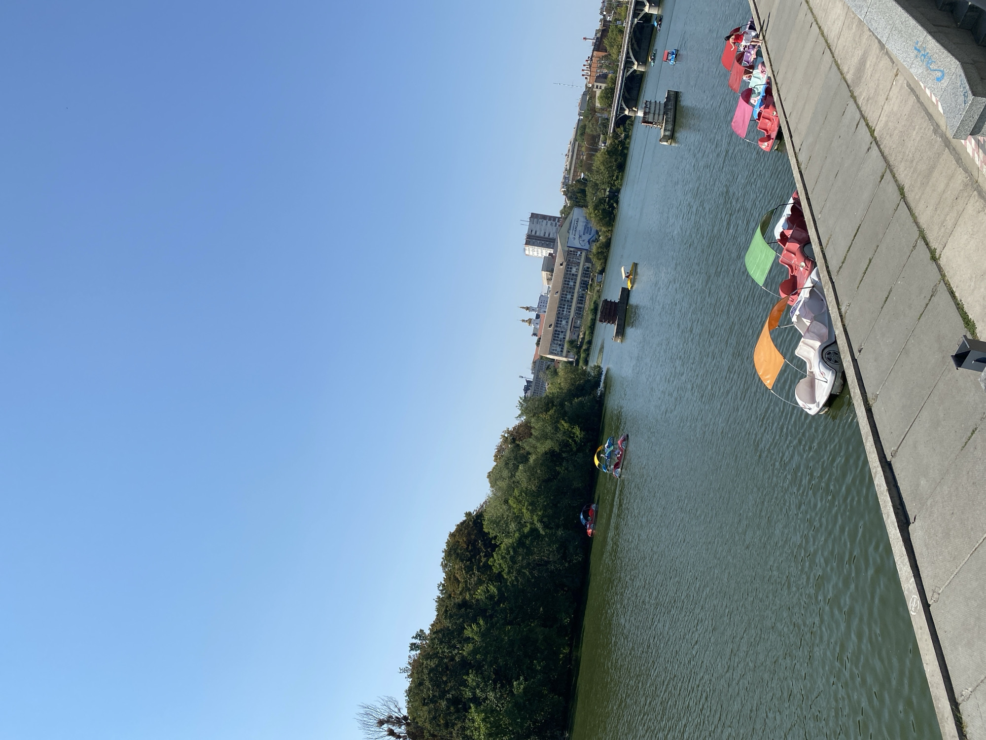 The image depicts a sunny day along the Southern Bug River in Vinnytsia, Ukraine. The foreground shows a concrete embankment with several colorful pedal boats docked along the edge. These boats are red, white, pink, and green, each with a canopy. Some of the boats are empty, while others appear to have passengers or are waiting to be rented.
In the middle ground, the river is calm and reflects the clear blue sky. Several more pedal boats are scattered across the water, with some people visible inside them, presumably pedaling. To the left of the river, a dense line of trees forms a lush green bank.
In the background, the city skyline is visible. Buildings of varying heights, including a tall, modern structure and some older, lower buildings with distinctive domed roofs, are present. A bridge crosses the river in the distance, with traffic visible on it. The overall scene suggests a recreational area in the city, likely a park or waterfront promenade.