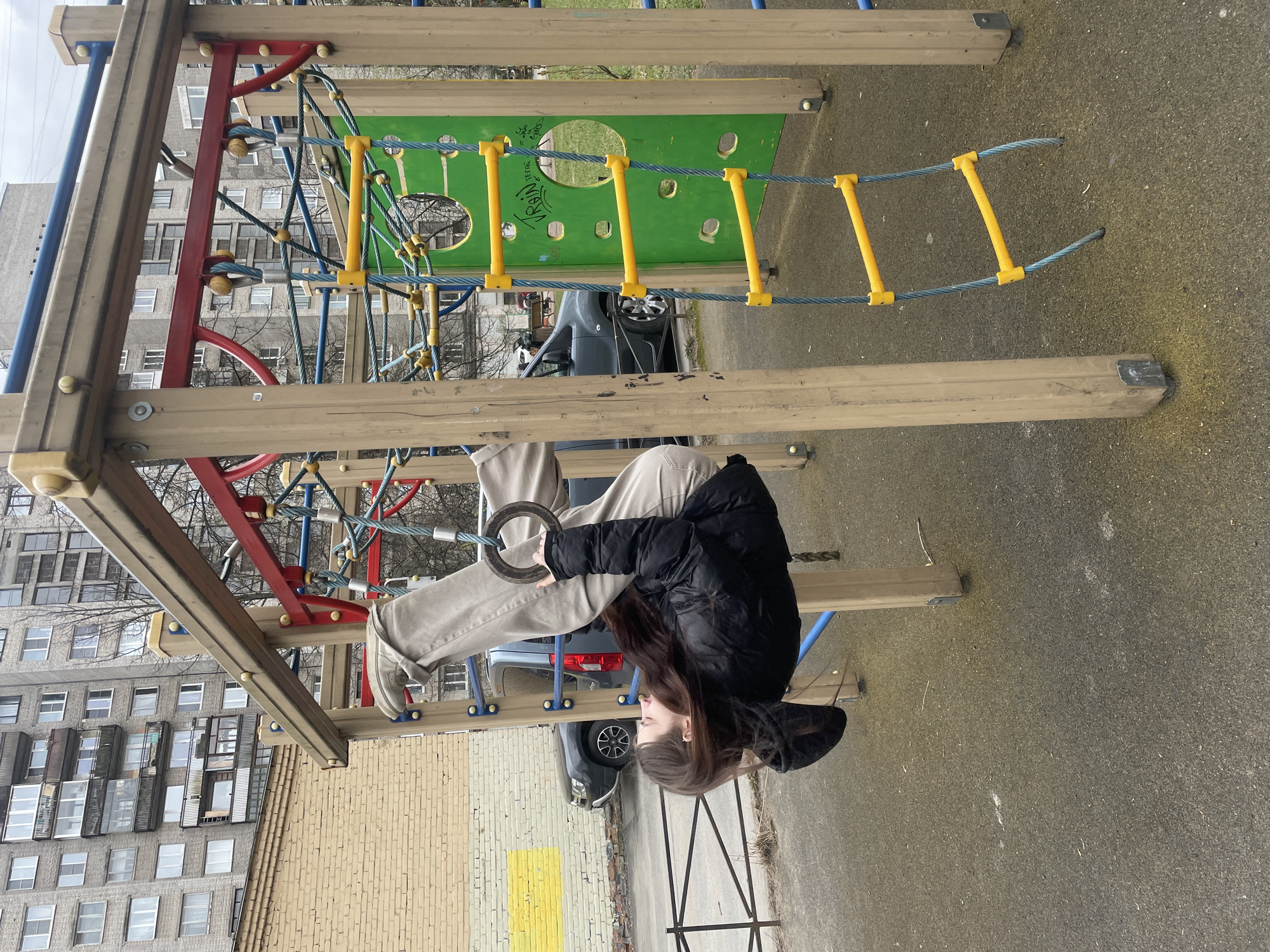 A young person, likely female, with long dark hair, is depicted hanging upside down from gymnastic rings on a playground. She is dressed in a black hooded puffer jacket, light beige trousers, and light-colored shoes, with her legs extended upwards. The gymnastic rings are attached to a colorful playground structure, which features sturdy wooden beams, red and blue metal supports, green plastic panels with circular cutouts, and yellow-runged rope ladders forming climbing nets.
The ground beneath the playground is a dark, paved surface, possibly asphalt or rubber matting. In the background, several large, multi-story residential buildings with numerous windows are visible, indicative of an urban setting. Leafless trees suggest a colder season, such as late autumn, winter, or early spring. A dark grey car is parked nearby.
The image is rotated 90 degrees clockwise, presenting the scene horizontally. The sky is overcast and grey, suggesting a non-sunny day. On one of the green plastic panels of the playground, graffiti is visible, partially reading "УКРАИ", which likely forms part of the word "УКРАИНА" (Ukraine). The scene captures a moment of playful activity in an urban environment, consistent with a public park or residential courtyard in Saint Petersburg, Russia.