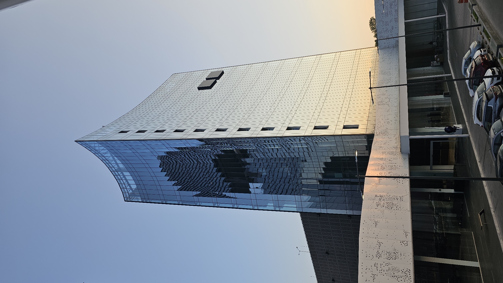 This image shows the architectural details of a modern building with a curved, glass-covered facade. The top portion of the building is constructed with a grid-like pattern of metal panels. There are dark, rectangular structures on the roof. The glass facade reflects the surrounding buildings. The building is located near a street with parked cars. The building's design includes a perforated white wall section. The sky is visible, ranging from a clear blue to a warmer yellow near the horizon, suggesting either sunrise or sunset. Based on visual clues, the building appears to be located in Puebla, Mexico.