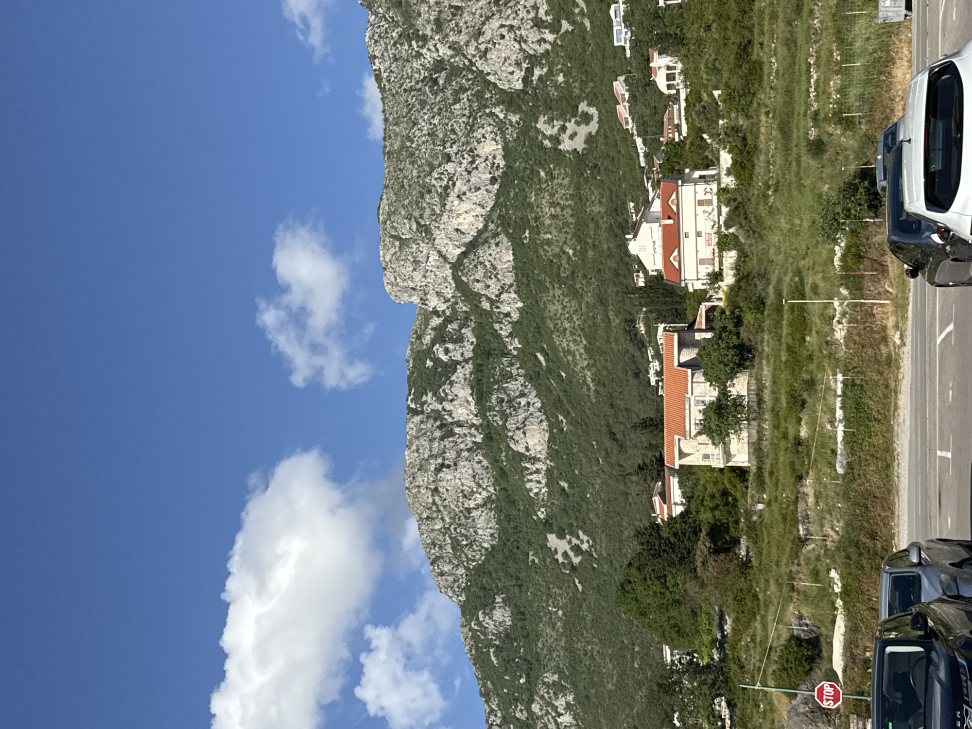 The image shows a view of the Rock of Gibraltar, in the city of Blizikuće, Montenegro. To the right of the rock, there are several houses and buildings. A road runs alongside the houses, with parked cars visible on the road. A stop sign is also visible on the right side of the road. The sky is blue with some white clouds.