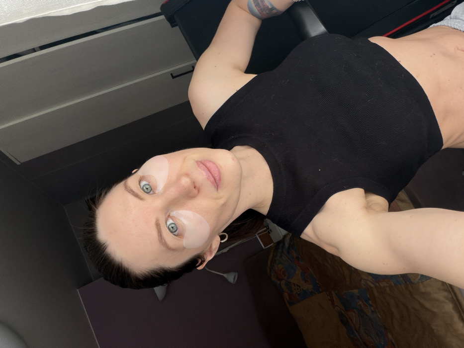A woman poses for a selfie, her face turned slightly to the side. She wears eye masks and a black tank top that shows a bit of her toned abdomen. Her eyes are a striking blue. Her left arm is visible, showing a colorful tattoo. The background includes a wall with drawers, a bed, and other furniture items. The picture has an unusual perspective, with the woman holding the camera at an angle.