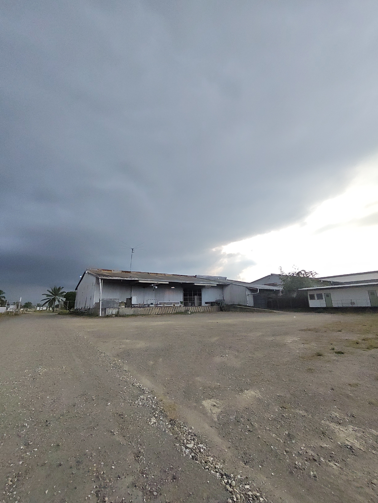 The image depicts an outdoor scene in Peurupok, Indonesia, under a dramatically overcast sky. The foreground is dominated by a rough, gravelly dirt path leading towards a long, low-slung industrial or warehouse-style building. This building is constructed of corrugated metal siding and has a weathered, rusty roof. There are several entrances or loading bays visible along its side, with some benches or seating areas outside. To the left of this main building, a palm tree stands out against the dark, heavy clouds. Further in the background, another smaller building with white walls and green trim is visible, suggesting an industrial or commercial area. The sky is a mixture of dark, ominous storm clouds on the left and brighter, lighter clouds on the right, with a sliver of sunlight breaking through. The overall impression is of a rural or semi-industrial location, possibly on the outskirts of a town, experiencing a change in weather. There are no people visible in the image.
